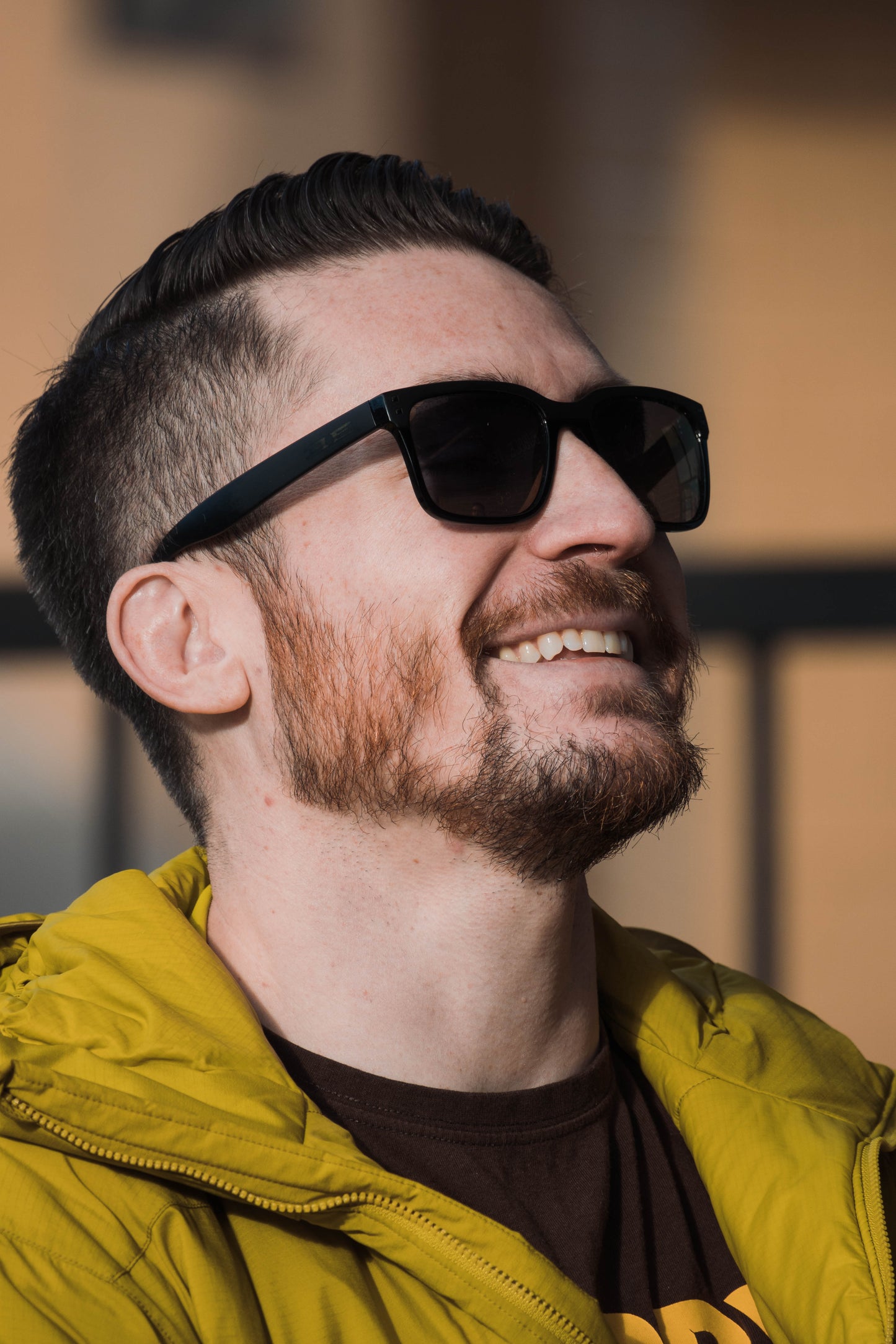 A man with short dark hair and a trimmed beard smiles outdoors, wearing Epoch Eyewear's Romeo sunglasses. He pairs them with a lime green jacket over a brown shirt, set against a blurred, warm-toned background.