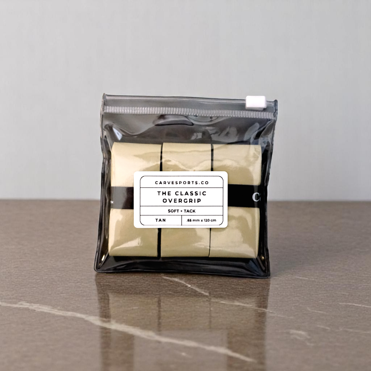 A small clear pouch with three tan Classic Overgrip tapes by Carve Sports (45 mm x 130 cm) rests on a smooth brown surface, set against a light gray wall.