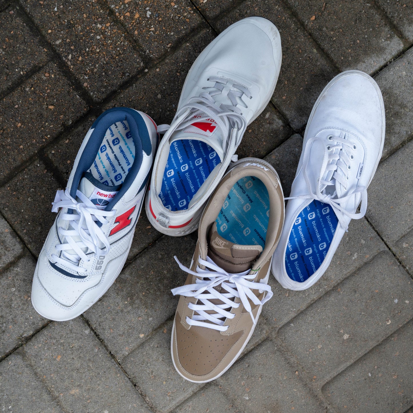 Four sneakers from Blumaka’s Recovery Bundle—two white, one tan with white, and one white with red and blue accents—are arranged on gray pavement. All feature Max Comfort Insoles with blue patterns for stylish support and all-day comfort.