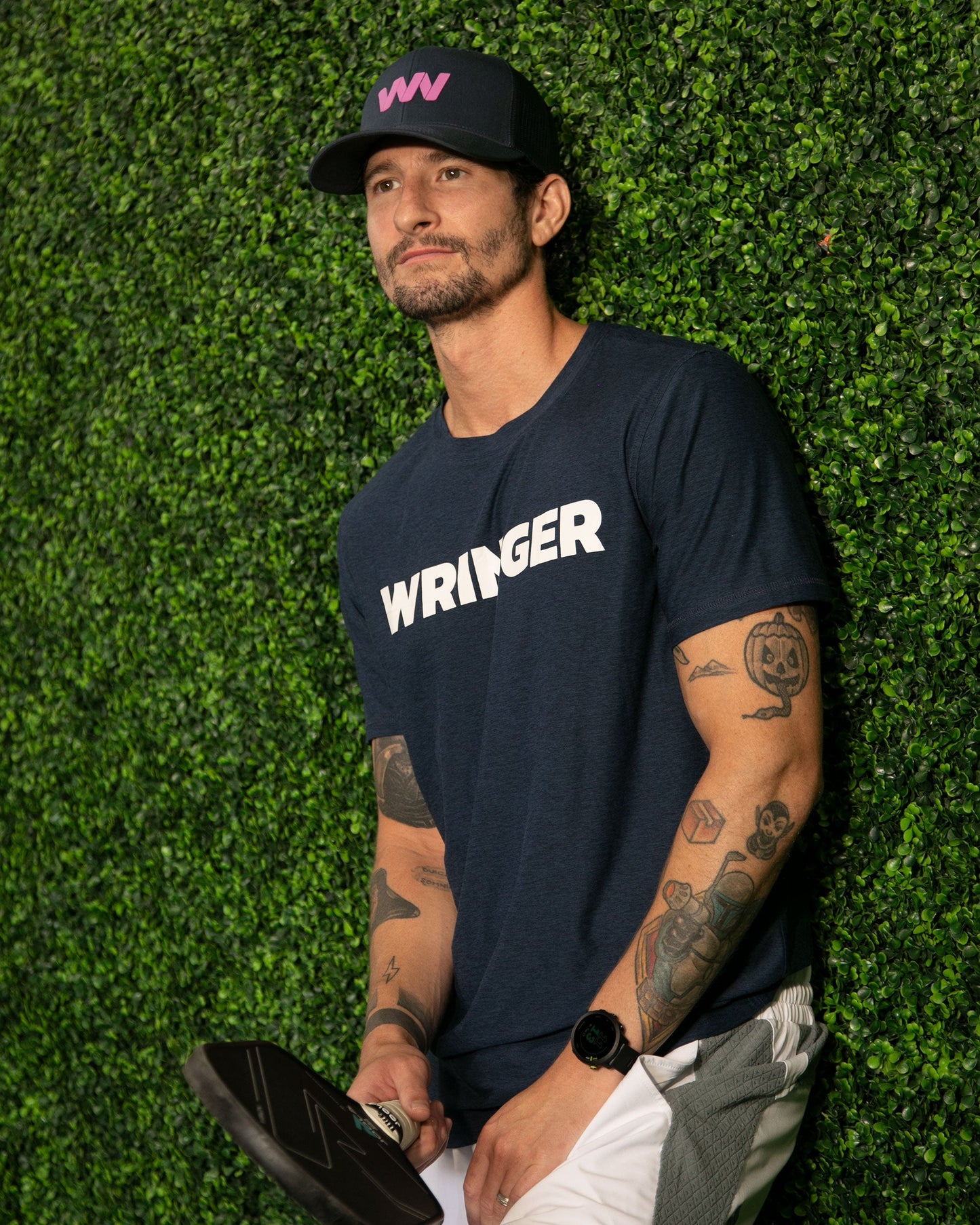 A tattooed, bearded man in a navy Men’s Short Sleeve WRINGER T-shirt by Wringer Wear and white shorts stands by a green leafy wall, holding a black paddle. He completes his athletic look with a navy cap featuring a pink logo, gazing to the side.