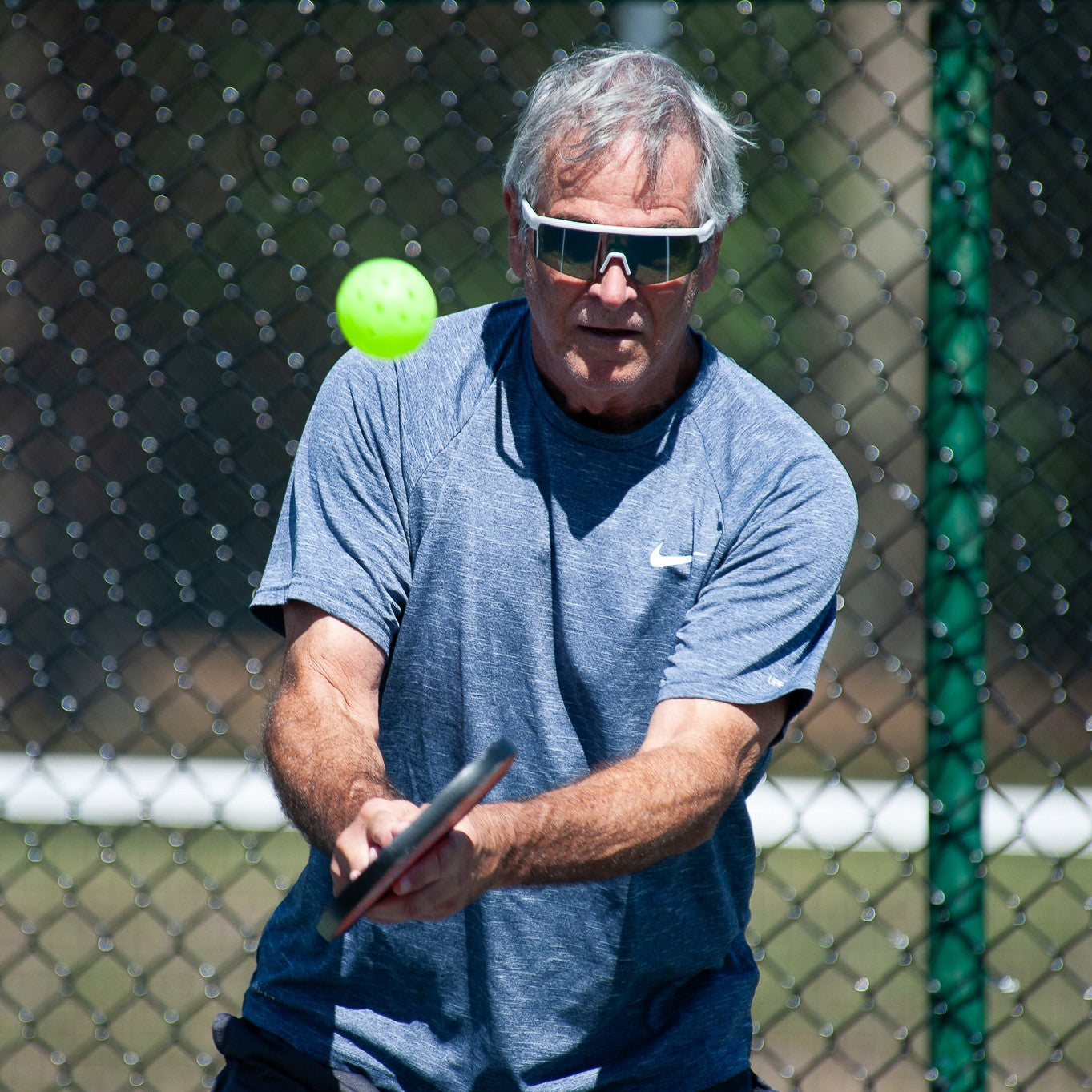 An older man wearing Dink Eyewear's Captiva White Pearl Pickleball Sunglasses plays pickleball outside in a blue athletic shirt, holding a paddle and bright green ball, with a chain-link fence and trees in the background.