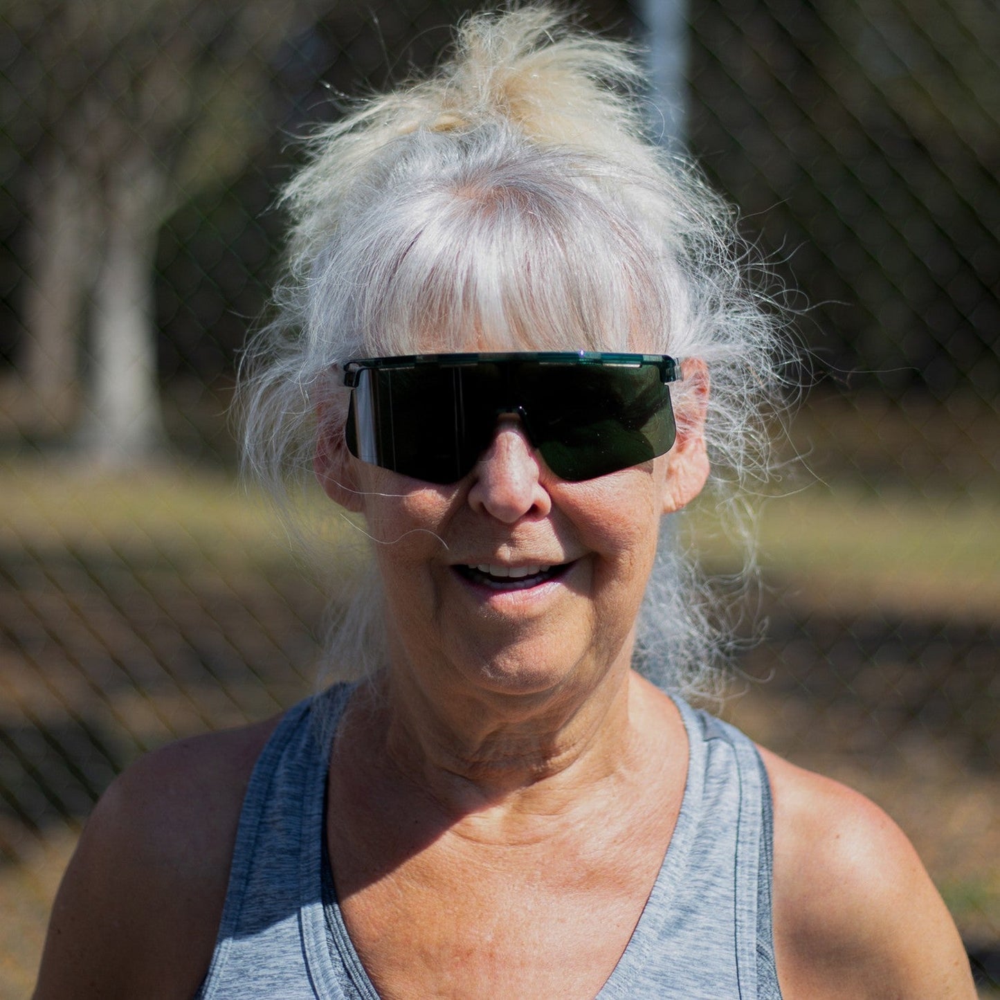 Older woman with long gray hair in a ponytail wears Dink Eyewear's Captiva Emerald Green Sunglasses and a gray tank top, smiling outdoors. The blurred background features a sunlit wire fence, trees, and grass.