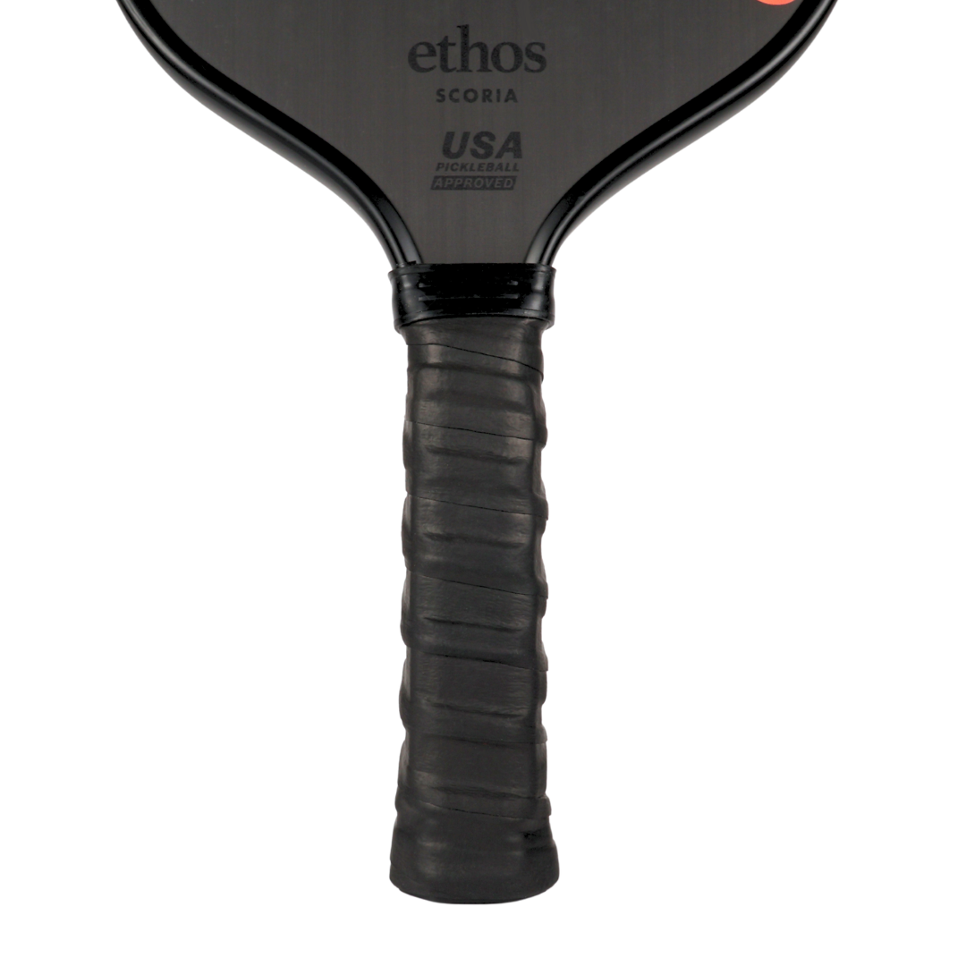 A close-up shows an Ethos Pickleball paddle handle wrapped with the Ethos Pro Undergrip, which reduces vibration for pain-free, confident play. "ethos SCORIA" and "USA PICKLEBALL APPROVED" appear in gray on the matte black face against a white background.