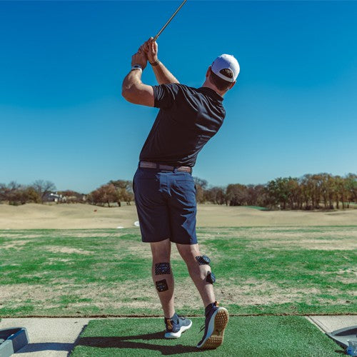 Wearing a black shirt, shorts, and a white cap, a man swings his club at the range. iReliev PRO Small Pads Refill Kit black tape supports his calf and knee muscles amid lush grass, blue sky, and trees in the background.