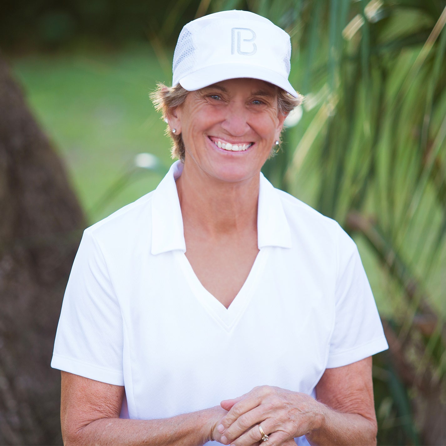 A smiling older woman with short hair and light skin stands outdoors among greenery, wearing a white Pickleball Bella PB Cap and a white collared shirt. Sunlight highlights her face as she clasps her hands together.