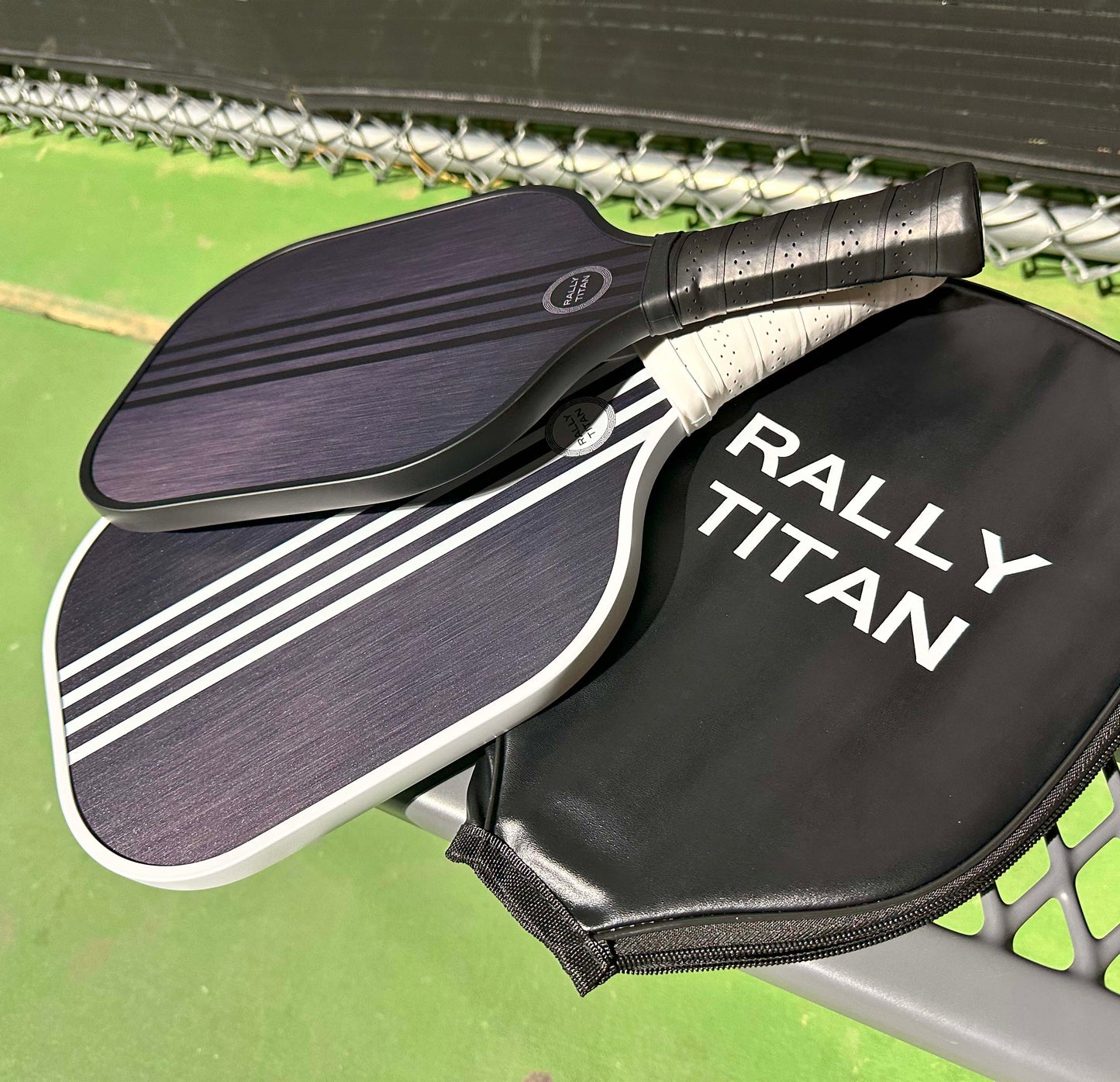 Two black Rally Titan Pickleball Aspro | 10MM paddles with T300 textured carbon fiber surfaces rest on a metal bench beside a matching case. The scene features a green court, chain-link fence, and dark barrier.