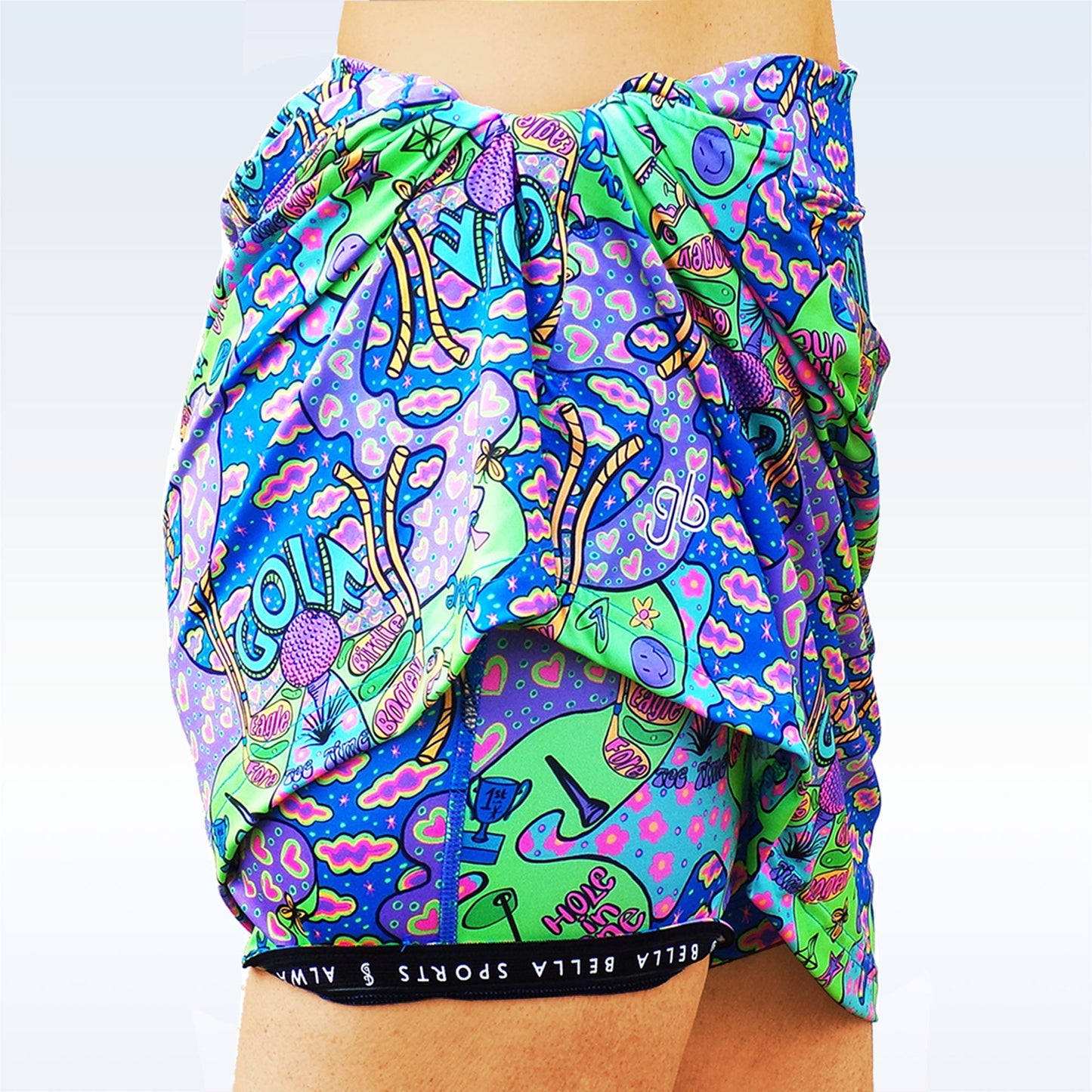 A close-up shows a person’s hip wearing the Pickleball Bella Groovy Straight Golf Skort by Pickleball Bella, featuring a colorful mushroom pattern and “BELLA SPORTS AWA” waistband, with a matching sarong draped nearby.