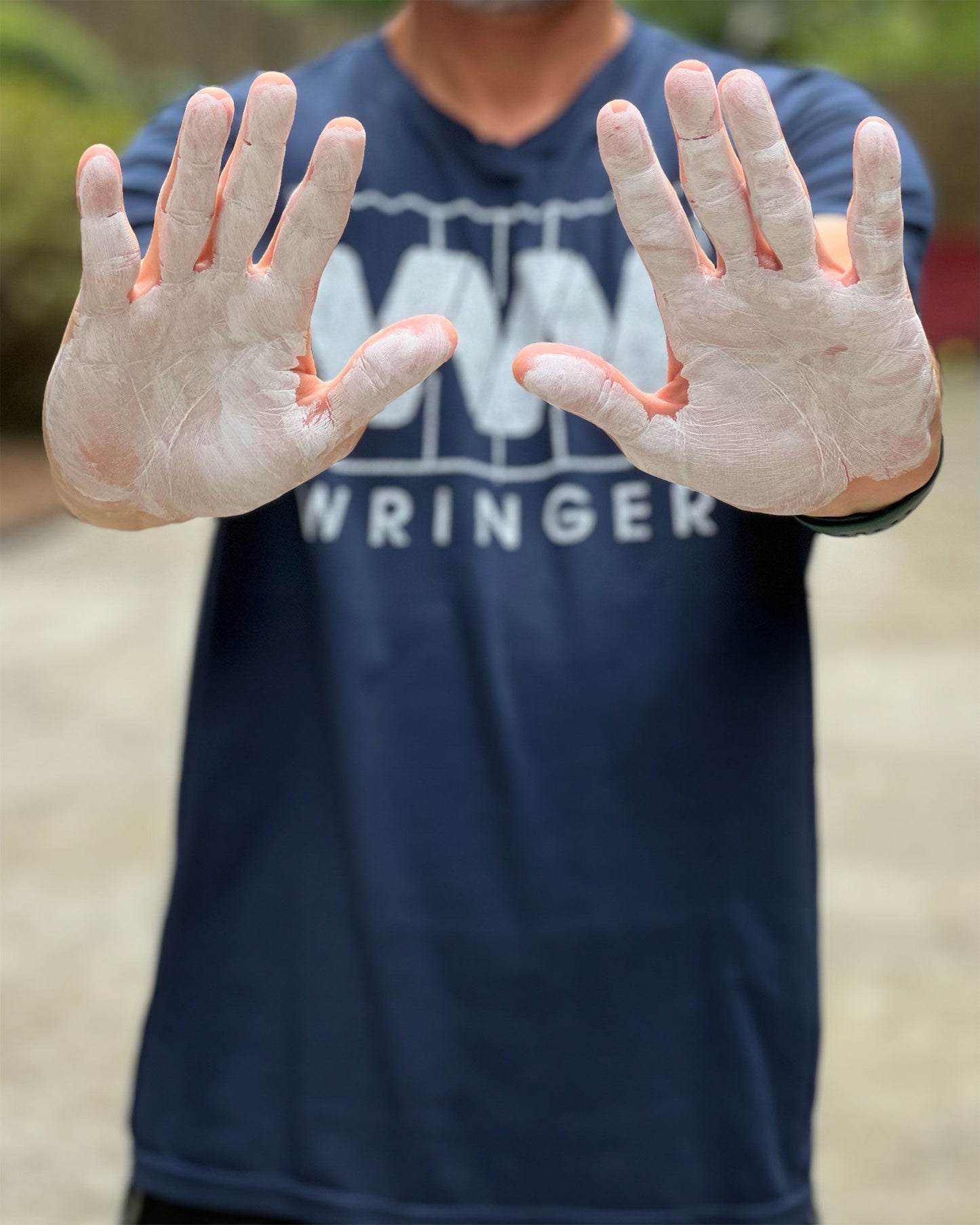 A person in a navy WRINGER WEAR T-shirt holds out chalk-covered hands, palms forward, showing off Wringer Wear Liquid Hand Chalk for dry grip—ideal for pickleball. A green, blurred background sets the scene.