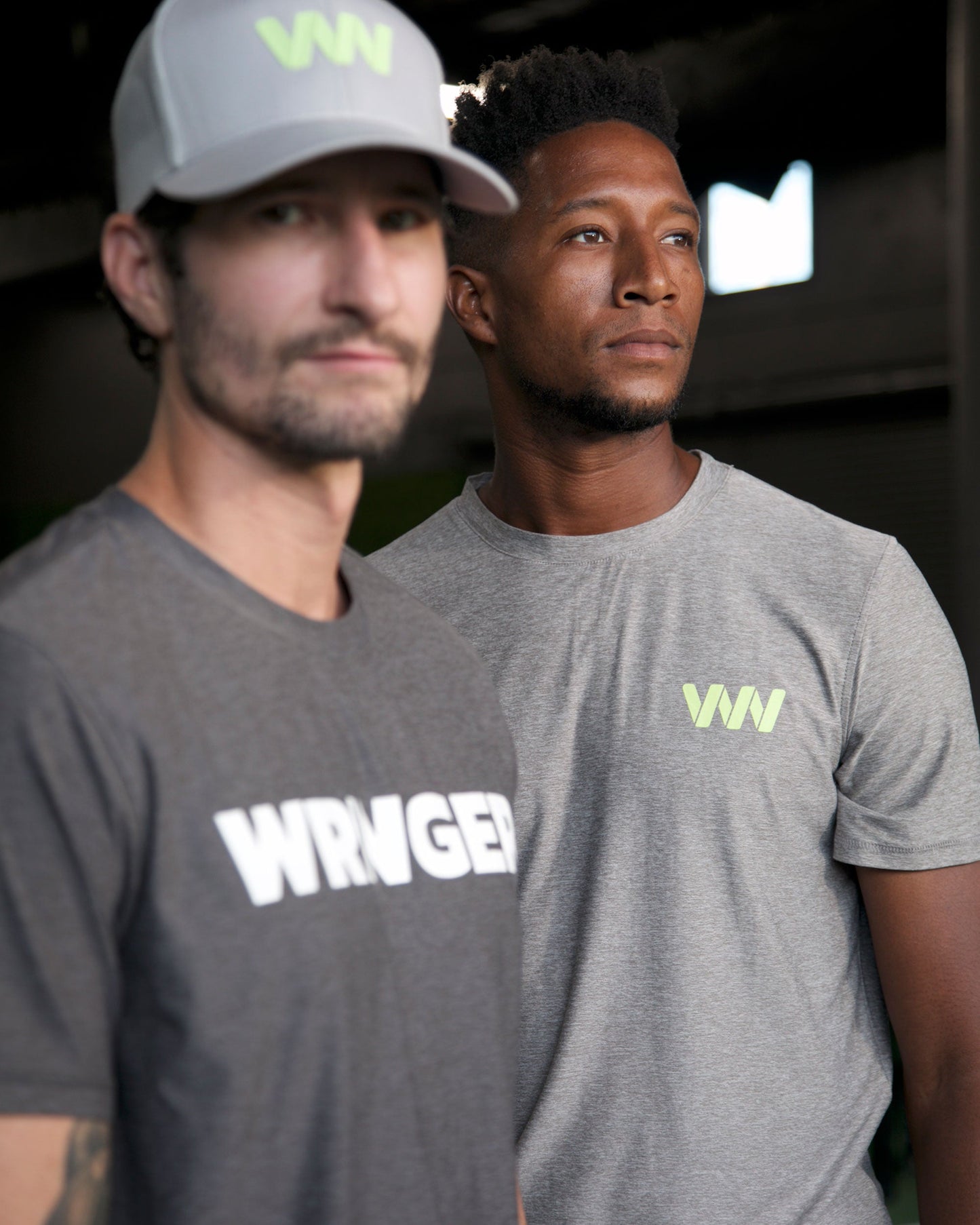 Two men indoors wear Wringer Wear Men's Short Sleeve Logo T-shirts in light gray—one with “WRNGED” in white, the other with a green WN logo. The left man pairs his tee with a matching cap. Both look right, highlighting the shirt's breathable comfort.