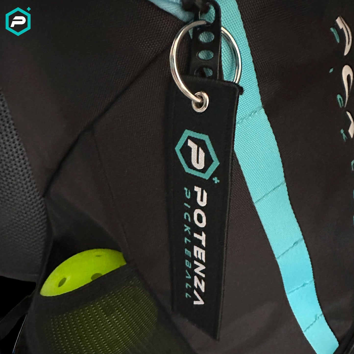 Close-up of a black bag with a turquoise stripe, featuring the Potenza Pickleball Logo Key Chain in a hexagon shape. A yellow-green pickleball with holes is partially visible in the mesh side pocket.