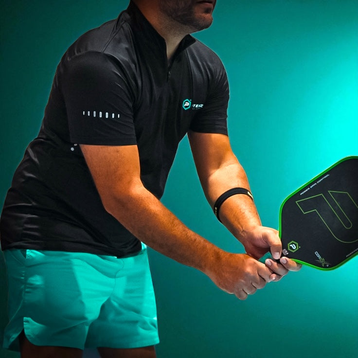 A man wearing the Potenza Pickleball Sport Athletic Polo (Quarter Zip Collar) in black and teal shorts holds a green and black paddle, ready to serve. The teal background matches his shorts, creating a vibrant, sporty vibe. His face is not visible.