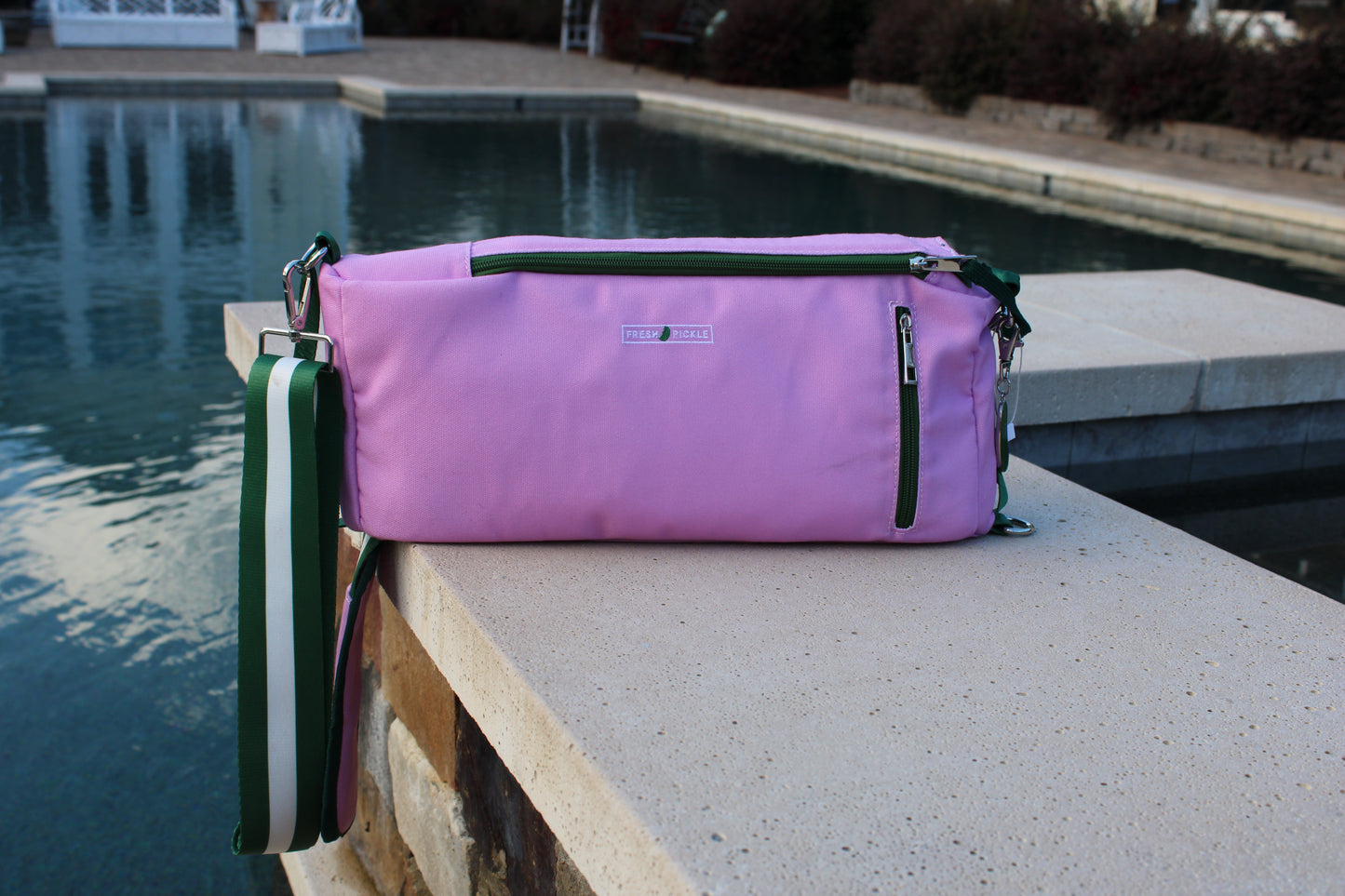A Fresh Pickle Insulated Cooler Bag in light purple, featuring a green zipper and a green-and-white striped shoulder strap, sits on a stone ledge beside a clear blue pool, with trees and shrubs behind it.