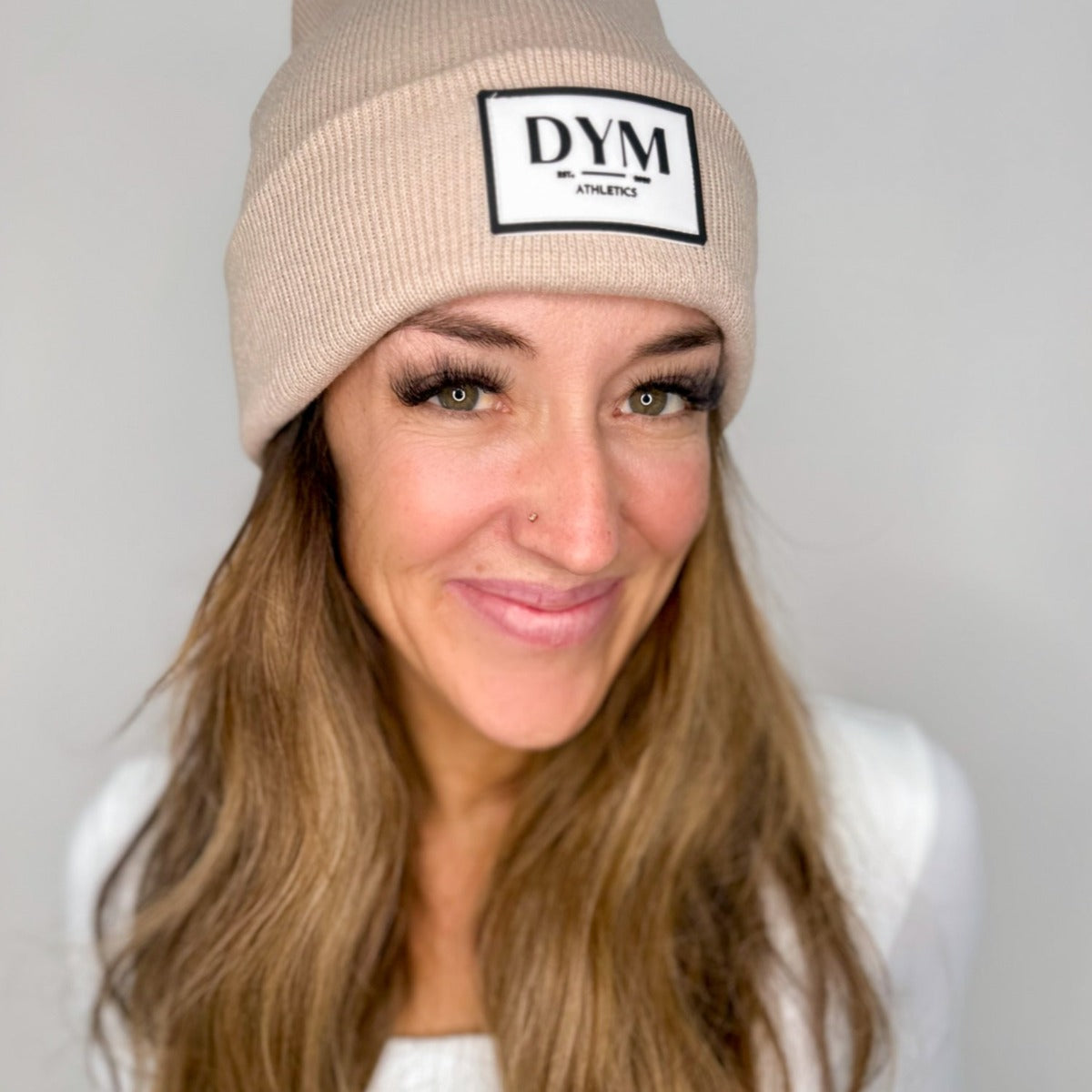 A woman with long light brown hair and a nose piercing smiles while wearing the DYM Beanie | SALE by DYM Athletics. She pairs it with a white top in front of a plain, light-colored background. Visible eyelashes add to her cozy look.