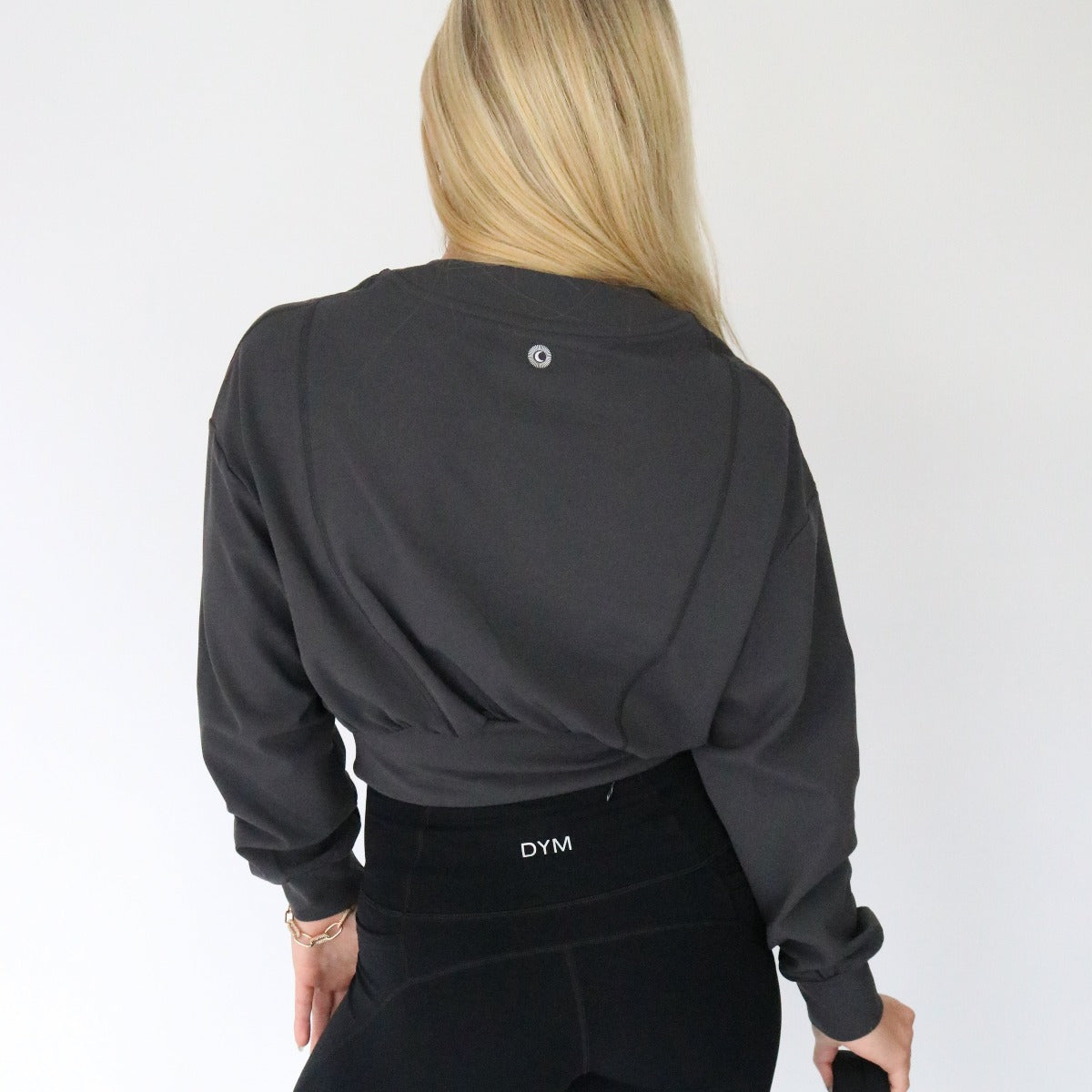 A person with long blonde hair, seen from behind, wears the Let's Brunch Bomber Jacket by DYM Athletics, channeling contemporary style against a plain white background while holding a small item in their left hand.