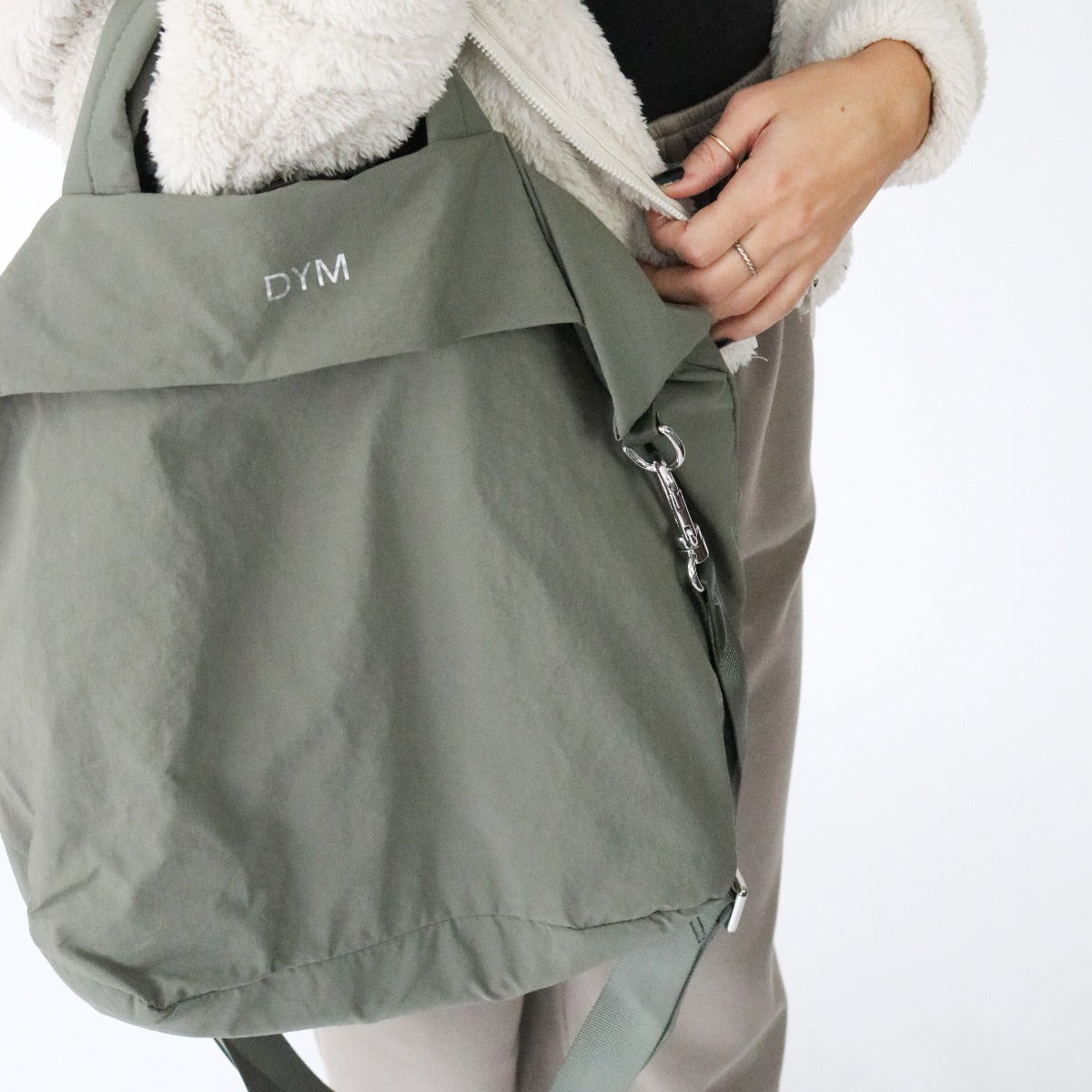 A person in a fuzzy beige jacket and taupe pants holds the DYM Athletics In Motion Bag, an olive green travel bag with white DYM embroidery. The photo shows only their torso, hand, and the bag against a plain white background.