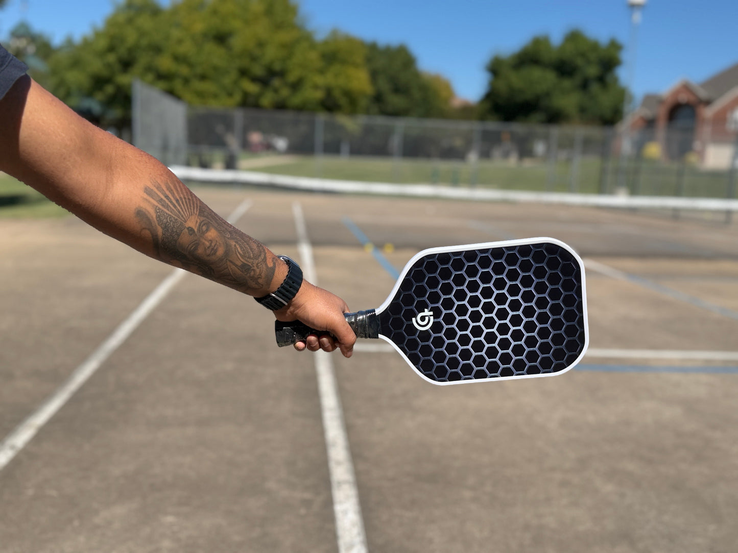 A tattooed arm holds out The Big Dink - Model 1 Pickleball Paddle, featuring a hexagonal face design, over an outdoor court with visible lines, fencing, trees, and houses in the background under a clear sky.