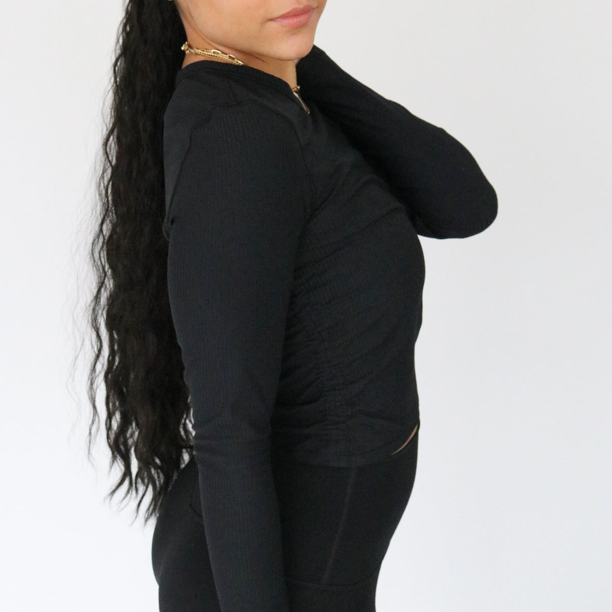 A woman with long, wavy black hair in a ponytail models the DYM Athletics Restore Ribbed Long Sleeve (SALE) with black pants, standing sideways and touching her neck against a plain white background.
