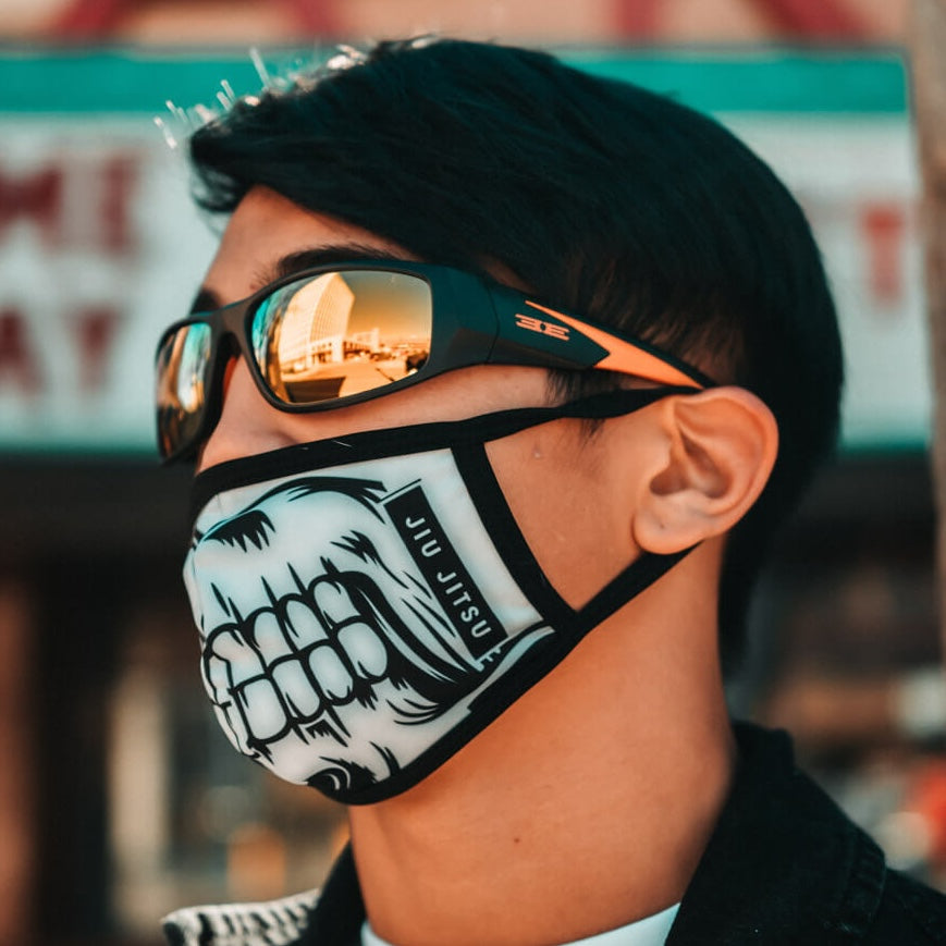 A person with short dark hair wears Epoch Eyewear’s Salerno full-frame sunglasses and a face mask with a grinning mouth graphic. The blurred background suggests an outdoor urban location with part of a sign in view.
