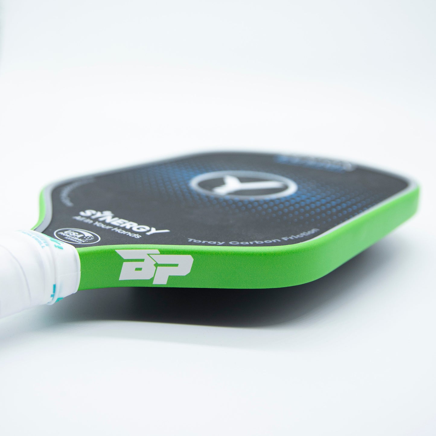 Close-up of a Bodhi Performance pickleball paddle with Premium Edge Tape - SOLIDS in green. The paddle features a black face, white handle, BP logo, “Synergy” text, and blue circles. The edge tape offers added protection. Background is white and blurred.