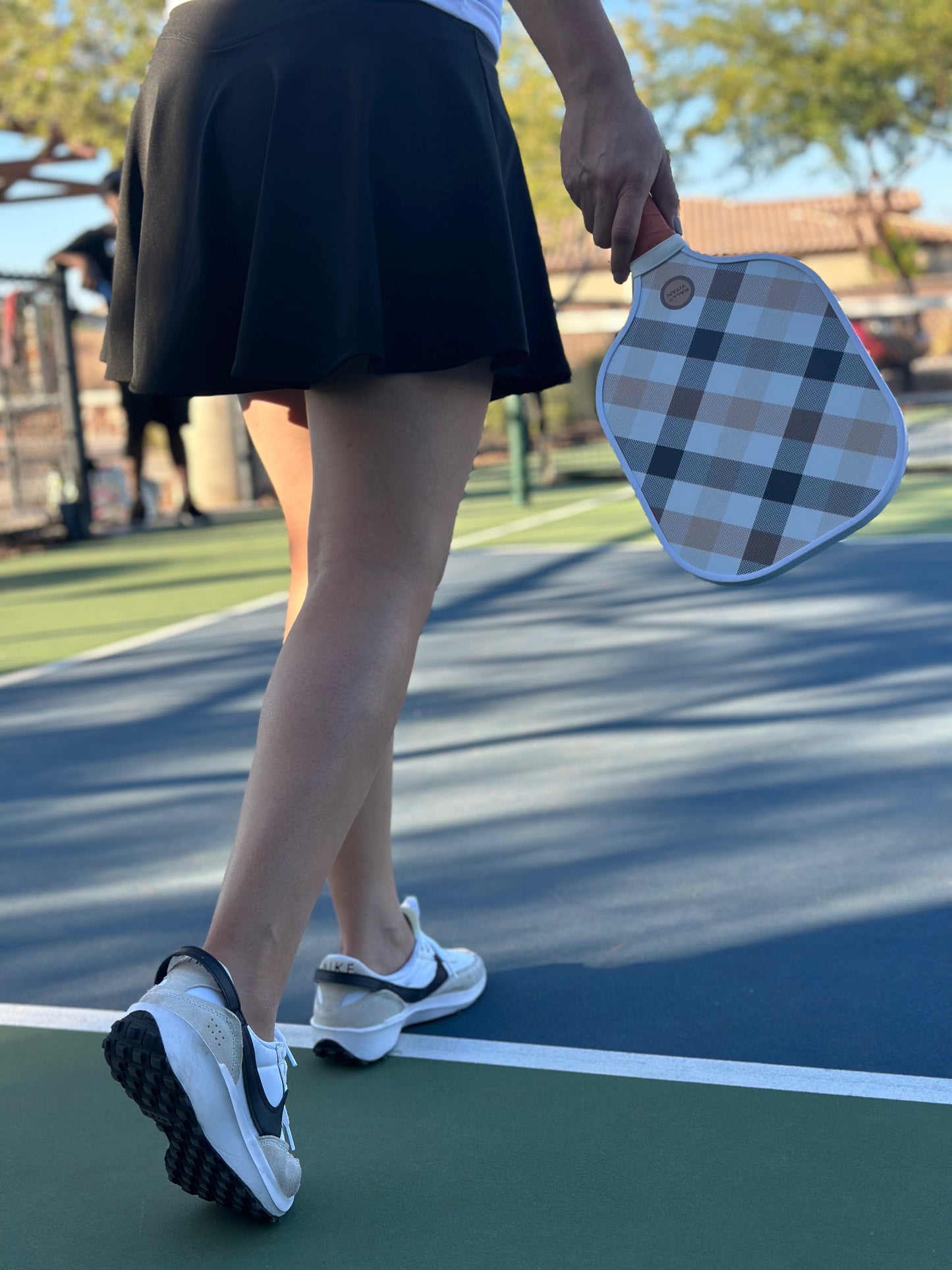 A woman on a pickleball court walks holding the Rally Titan Pickleball Plaid Luxe paddle with a 10MM T300 textured carbon fiber surface. She wears a black tennis skirt and white top; the background features blue and green court lines and trees.