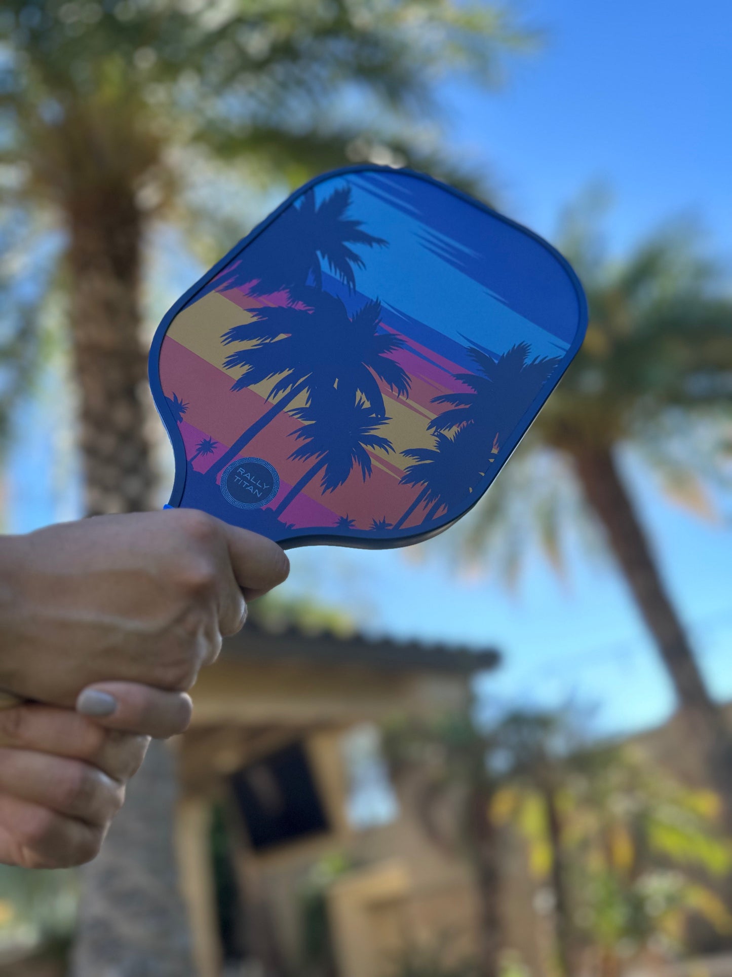 A hand holds the Rally Titan Pickleball Palm Beach paddle, featuring a 10MM T300 textured carbon fiber surface and a tropical sunset with palm trees, perfect for summery outdoor games.