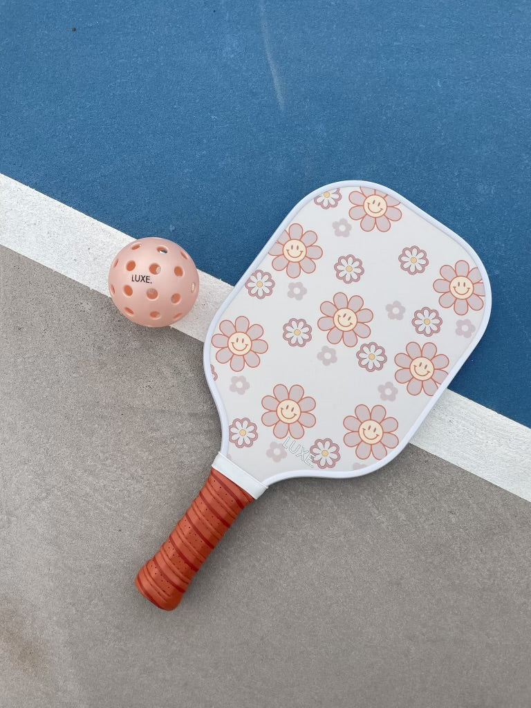 The LUXE Pickleball Smiley Flower paddle, USA Pickleball Approved, features a red handle and a white face with pink and orange flowers. It rests beside a pink LUXE ball on the court divided by a white line—perfect for beginners.