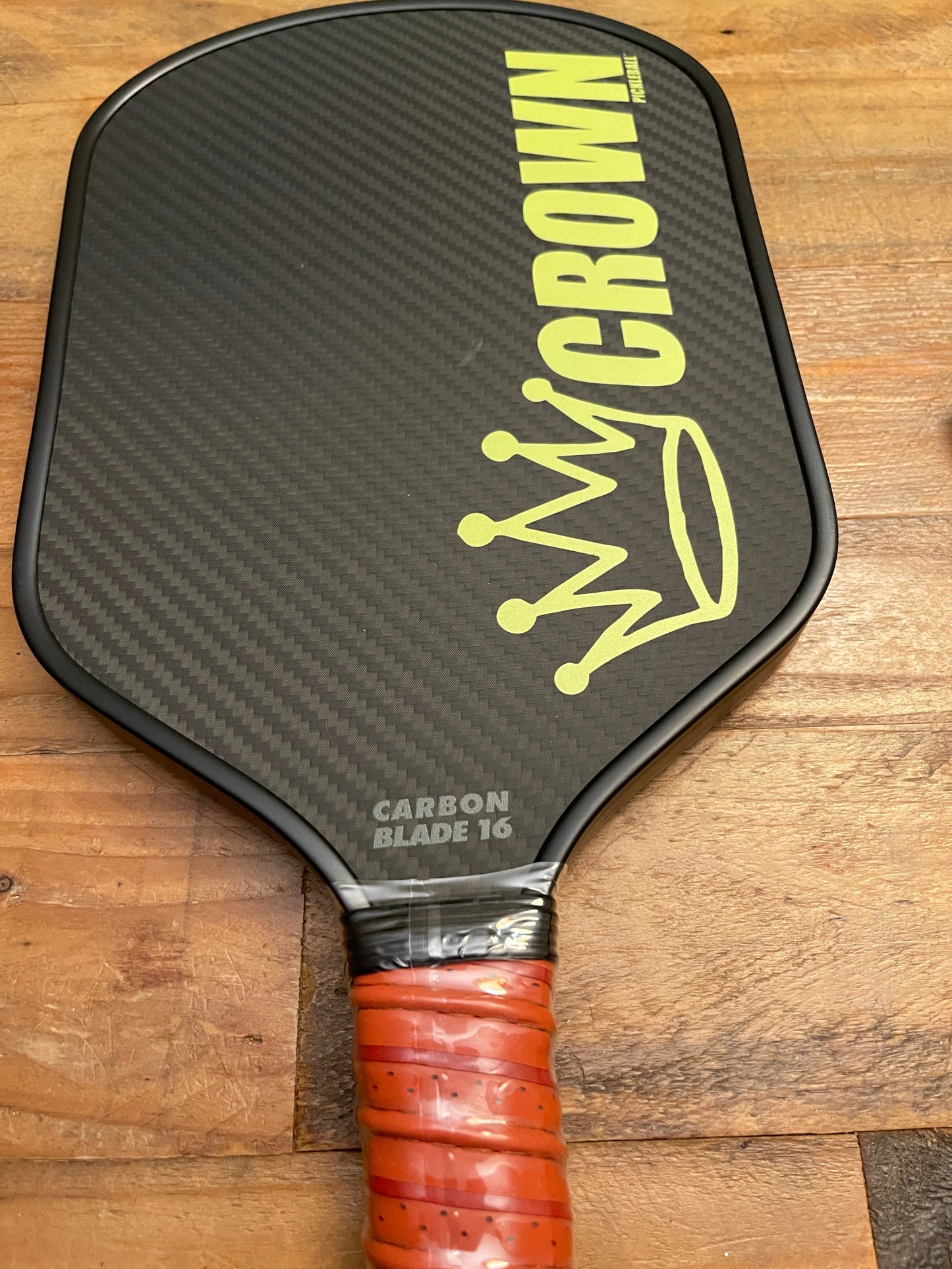 A CROWN PICKLEBALL - Carbon Blade 16 (BLACK Paddle) by Crown Pickleball with visible carbon fiber texture, "CROWN" and a yellow crown logo, plus an orange grip tape handle rests on a wooden surface.