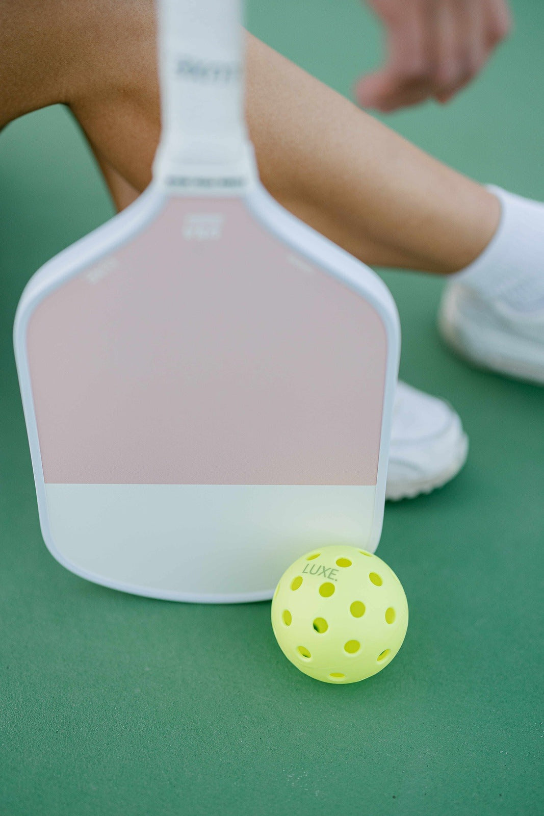 A close-up of the LUXE Pickleball Dinker carbon fiber paddle and a yellow pickleball on a green court. In the background, only the lower legs and hand of a person in white athletic shoes and socks are visible as they sit.