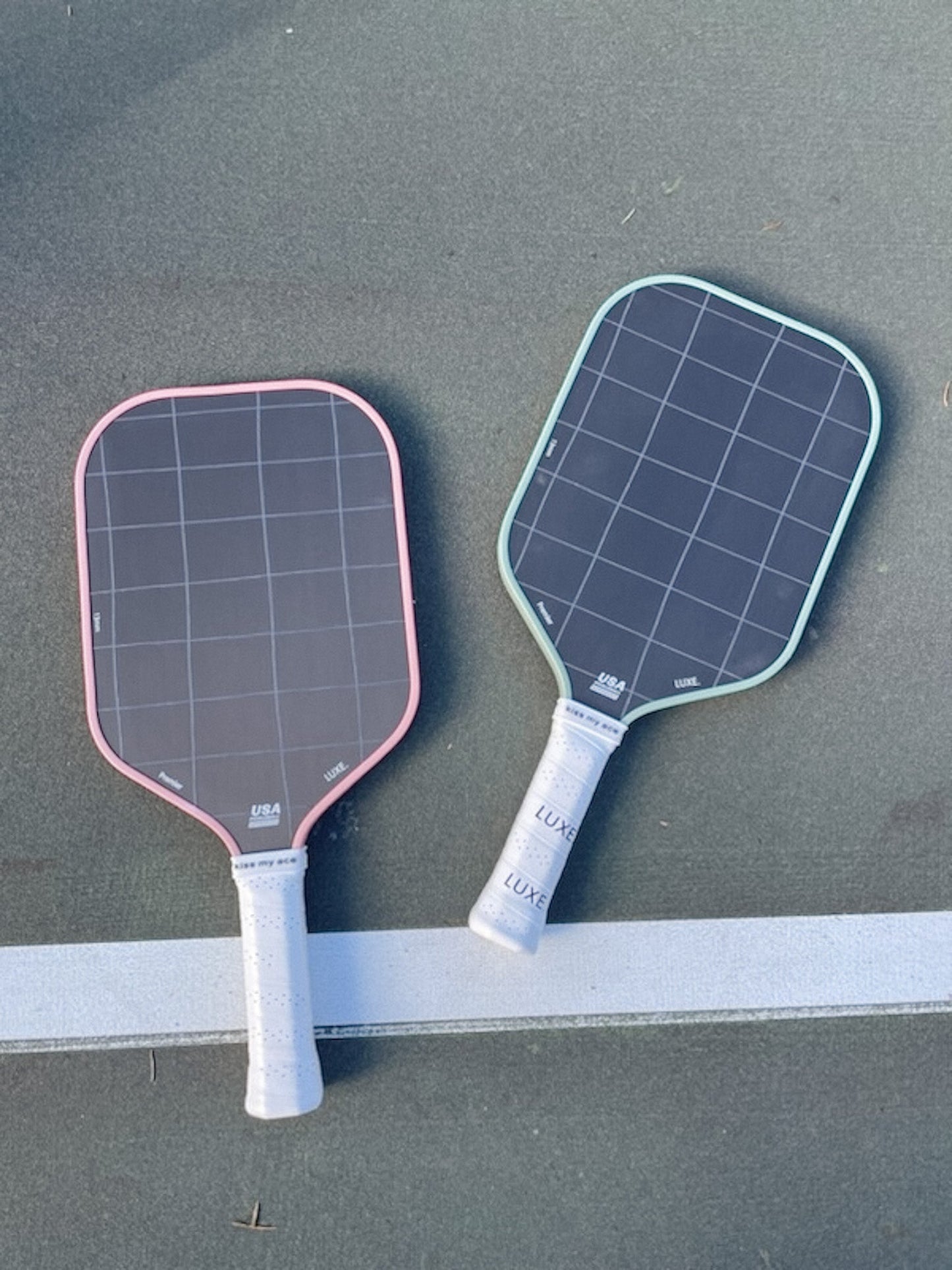 Two LUXE Pickleball Premier paddles with white handles and Toray t700 carbon fiber construction lie side by side on an outdoor court. One has a pink border, the other teal; both display a black grid pattern above a white line.