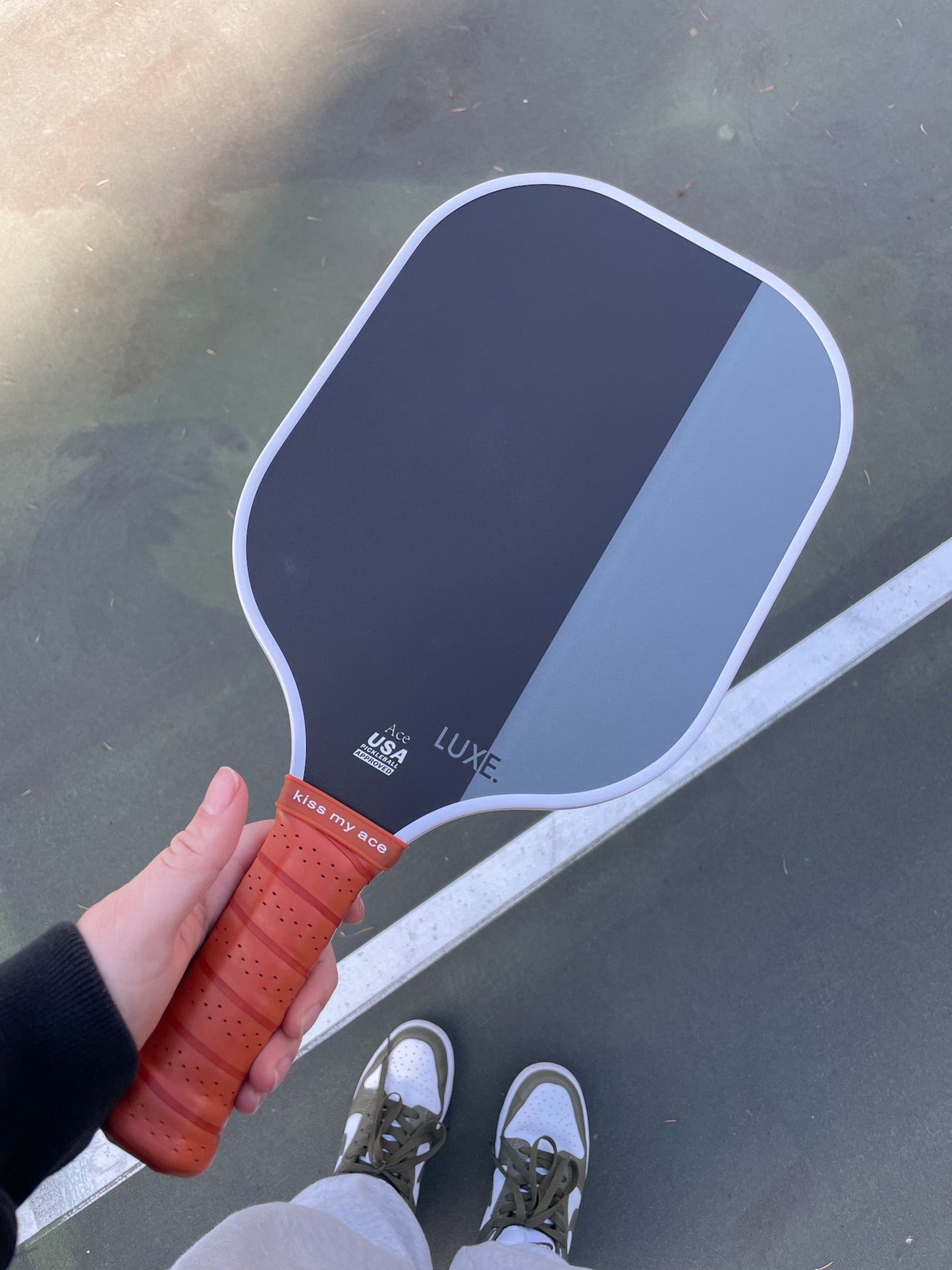 A person holds the LUXE Pickleball Dipped - Ace paddle, featuring a split black and gray face with "LUXE" branding and a brown grip reading "KISS MY ACE." Distinct colors pop against the court, with shoes, pants, and sweatshirt partially visible behind.