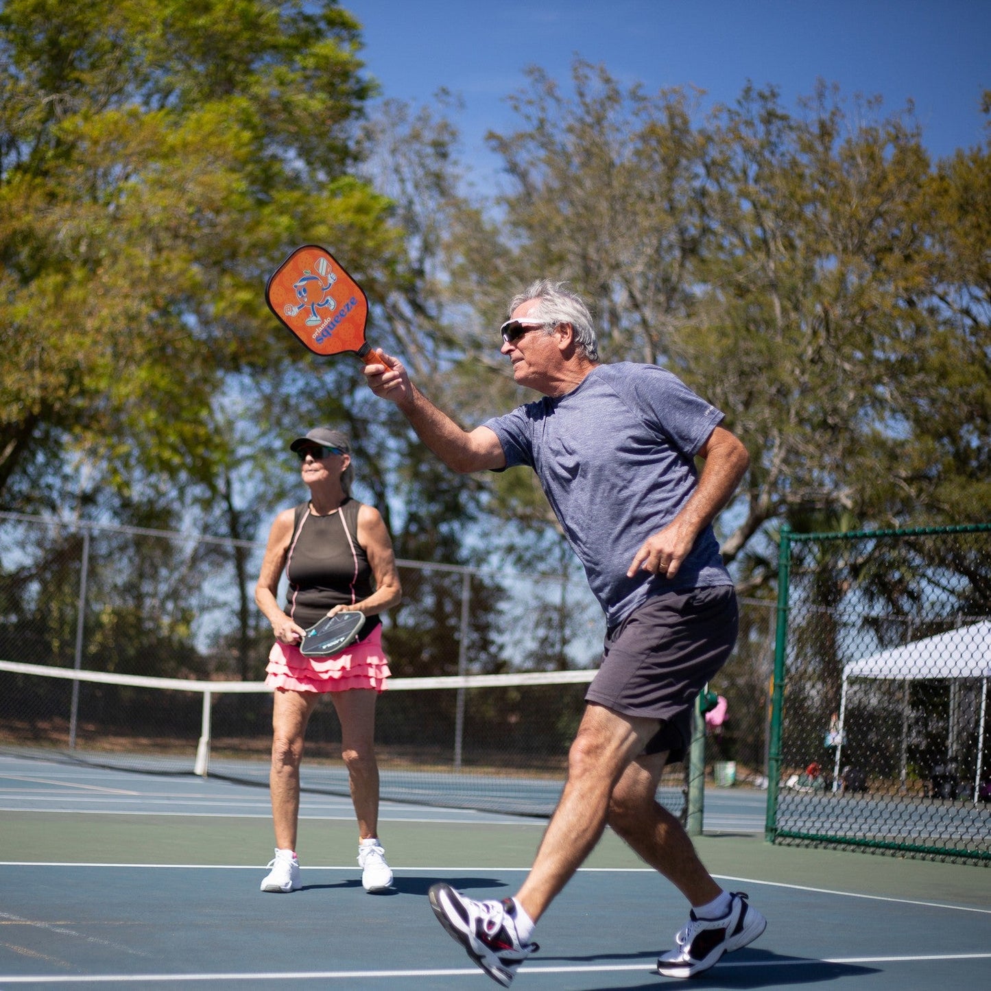 Two older adults play pickleball outdoors, both wearing Dink Eyewear Captiva Large Pink Pearl Sunglasses. The man in a gray shirt swings his paddle, while the woman sports a black tank top and pink skirt. Trees and a fence frame the action.
