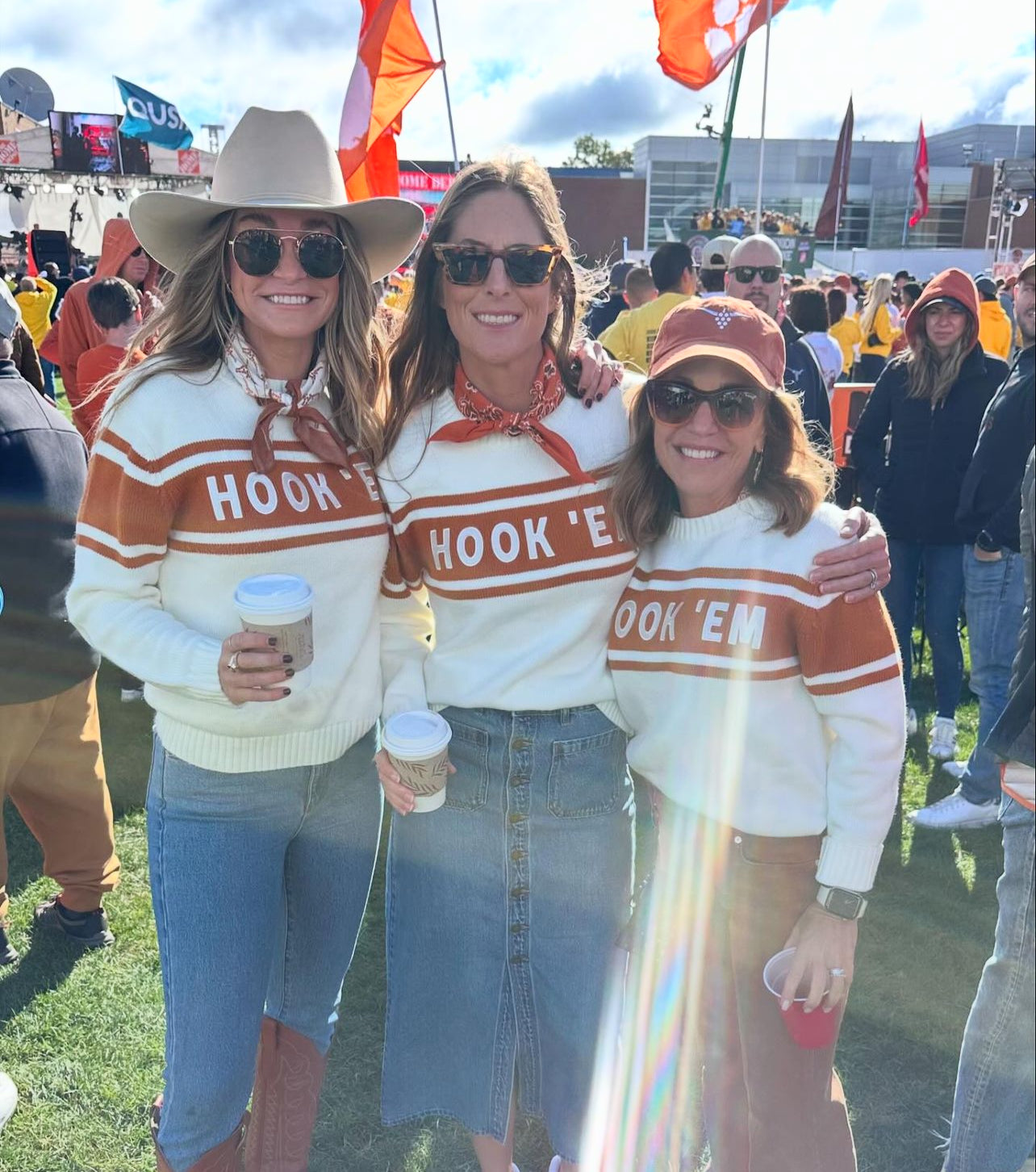 Three women beam outdoors at a sports tailgate, showing off their Runway Athletics HOOK 'EM Tennis Sweater - Limited Edition in white. Paired with denim and sunglasses, they display Longhorn pride as orange flags wave behind them under a partly cloudy sky.
