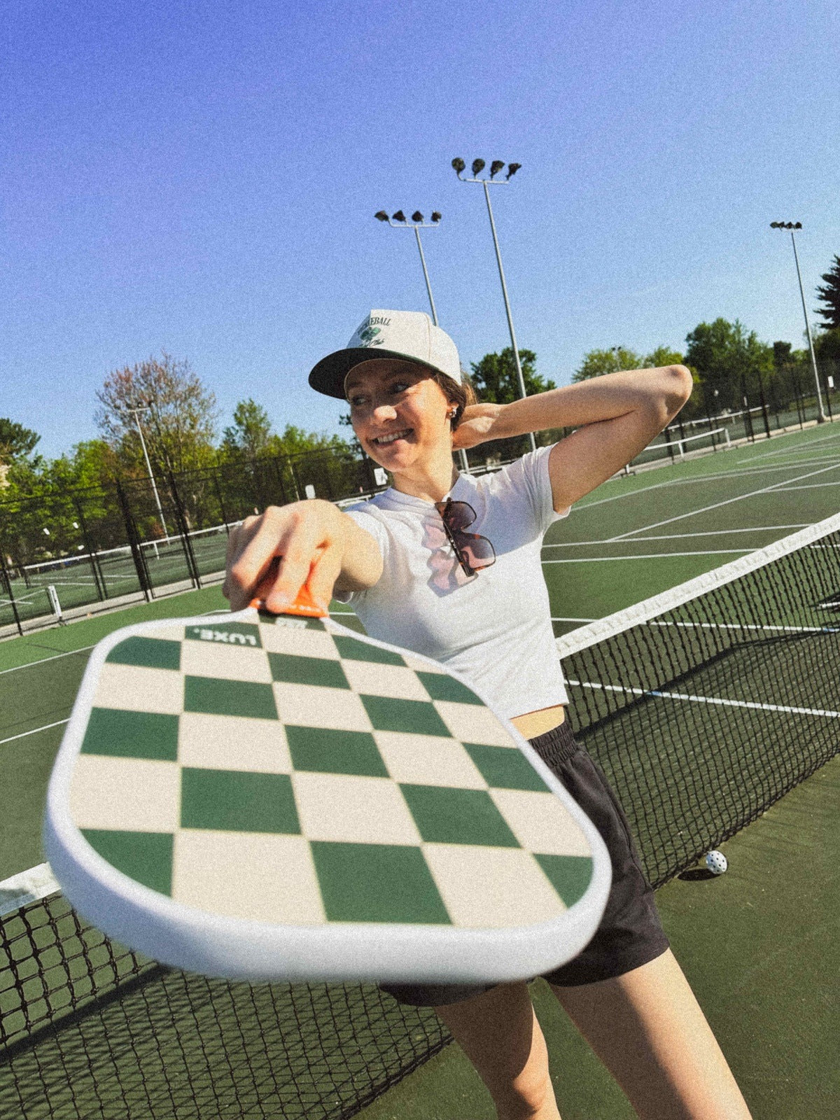 A smiling woman in a cap and sunglasses holds a LUXE Pickleball Checkmate paddle toward the camera on a sunny outdoor court, wearing a white shirt and black shorts, with green trees, nets, and lights under the blue sky behind her.