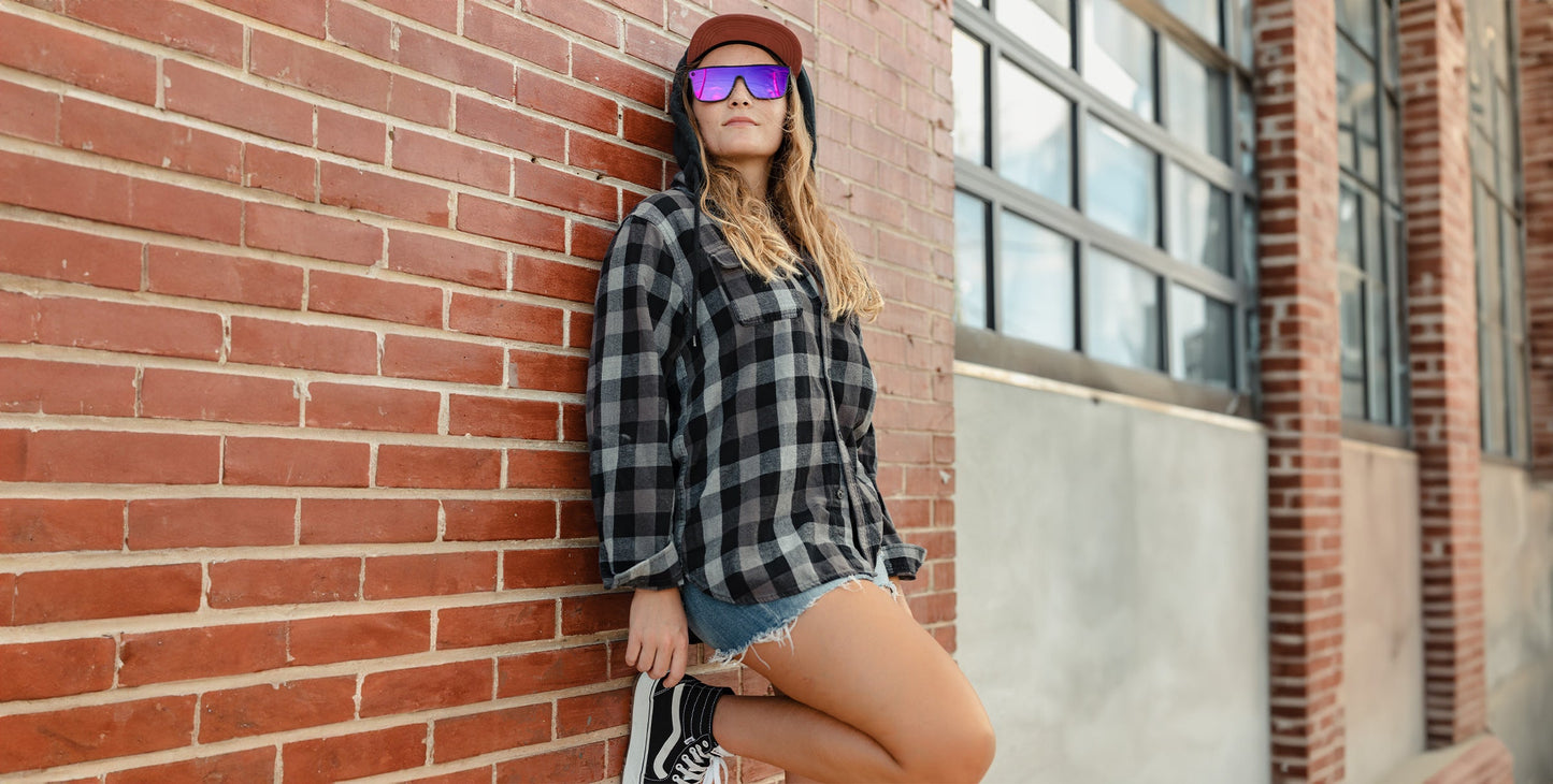 A person in a plaid shirt, denim shorts, and sneakers leans against a brick wall wearing Neven Eyewear's 53X polarized sunglasses with UV400 protection. They sport a cap, have long hair, and large windows reflect the sky beside them.