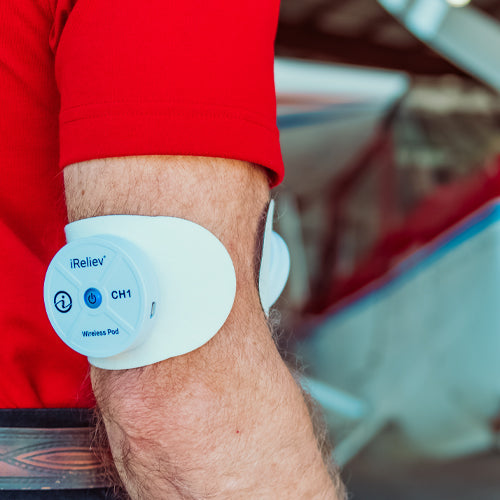 A person in a red shirt uses the iReliev Wireless Large and Small Electrode Pads Refill Kit on their upper arm; the white device with blue icon buttons labeled CH1 is attached, with a blurred airplane and hangar in the background.