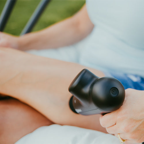 Seated on a white cushion, a person in a light blue shirt and denim shorts uses the iReliev Percussion Massage Gun, firmly massaging their thigh to promote blood flow. Their other leg appears partially in the background.