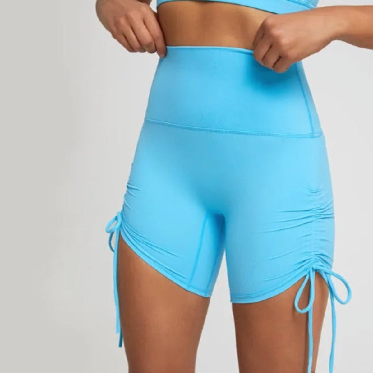 A person wears DYM Athletics' AIRE Ultra-Soft Ruched Biker Shorts in bright blue, featuring high waist, adjustable drawstrings, and ruched sides, paired with a matching crop top. Only their torso and upper legs are visible against a plain background.