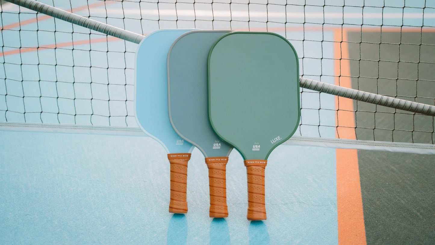 Three LUXE Pickleball Core Collection paddles with brown grips—two light blue, one green—lean upright against a net on a blue and black court with orange lines. Each USA Pickleball Approved paddle has a textured surface and partially overlapping faces.