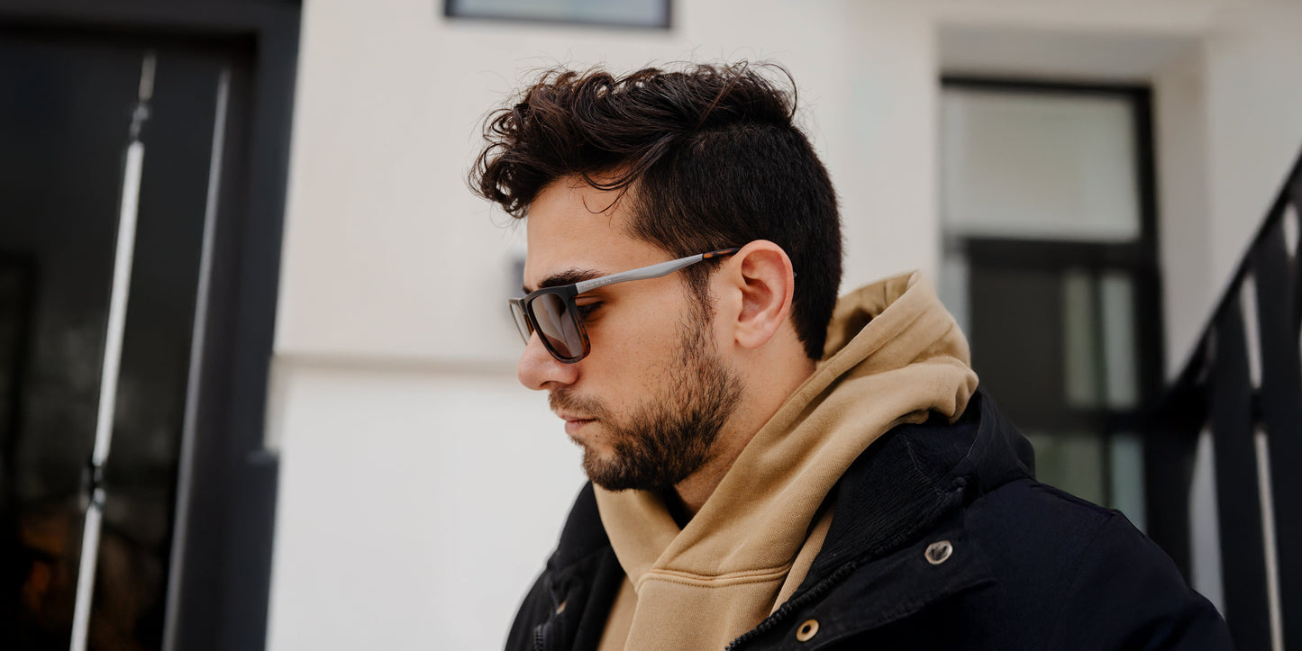 A young man with dark hair and a beard, wearing Neven Eyewear's Repeater polarized rectangular sunglasses, a tan hoodie, and a black jacket, stands outside in front of a modern white building, looking down seriously.