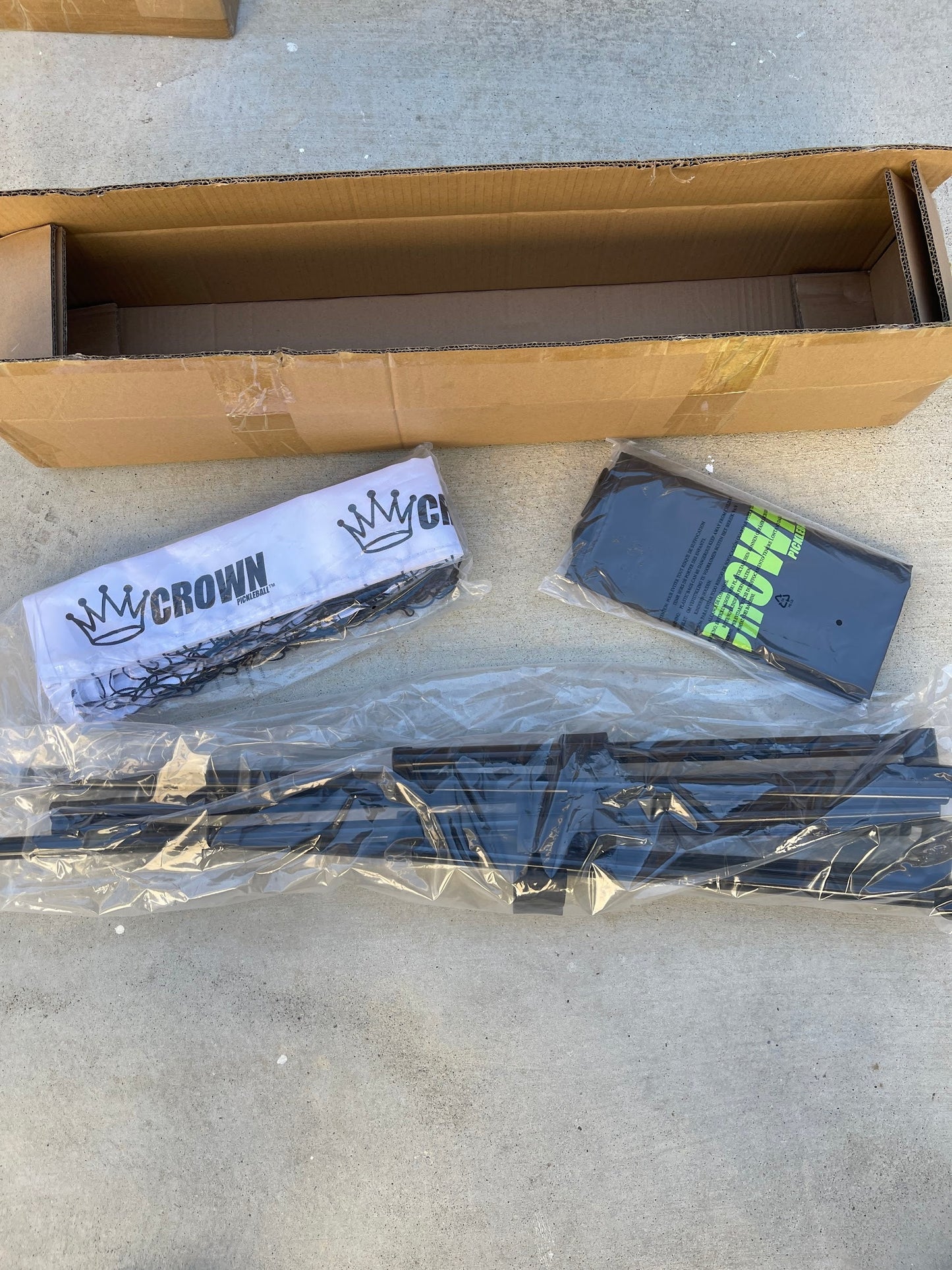 A CROWN PICKLEBALL - Full-Size Net (White/Black) by Crown Pickleball sits open on concrete, showing a branded white cloth, a black piece with green text, and several black metal bars in clear plastic for the portable net.