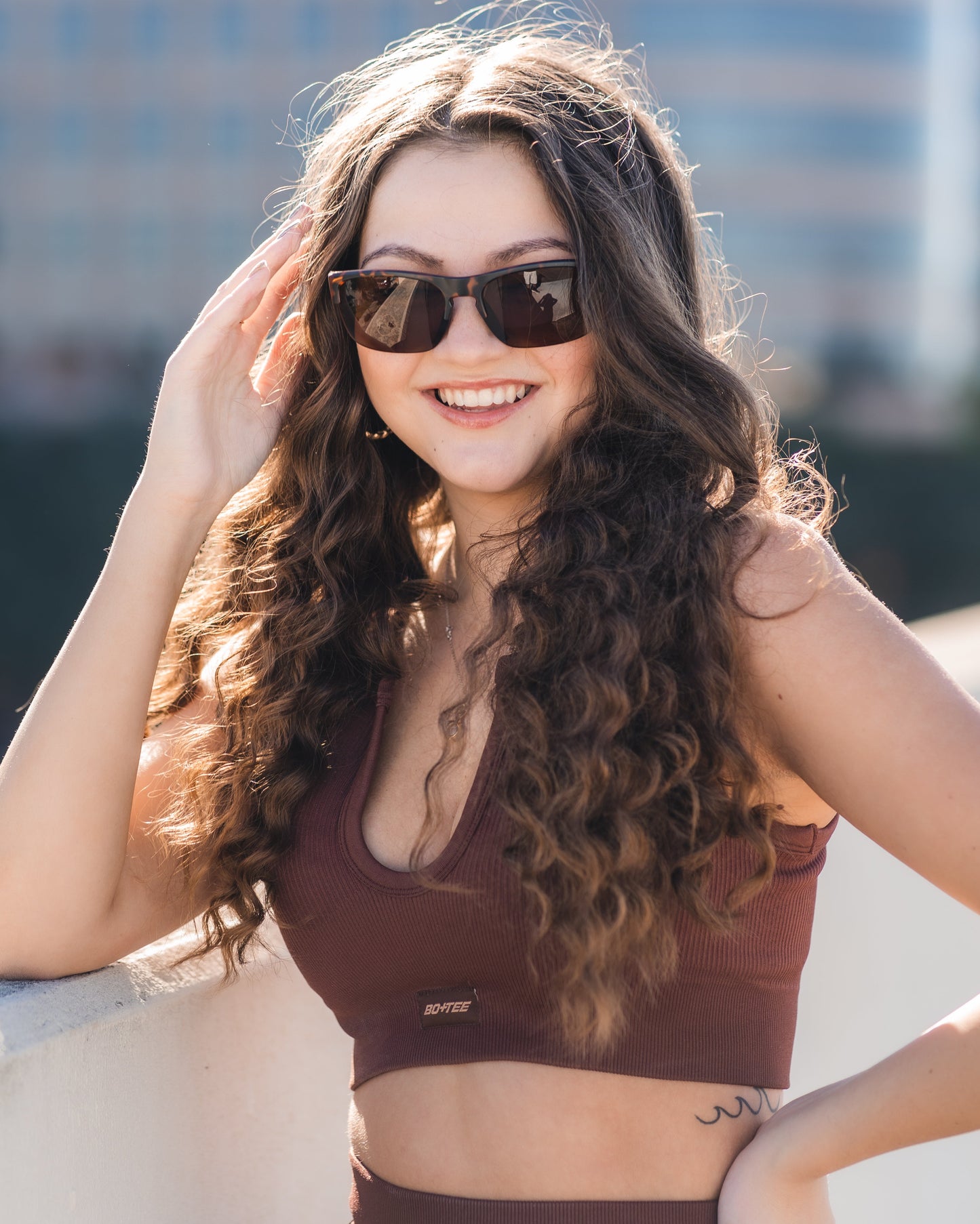 A woman with long wavy brown hair smiles outdoors in sunlight, wearing stylish Victor Sunglasses by Epoch Eyewear and a burgundy crop top, with blurred buildings and greenery in the background.