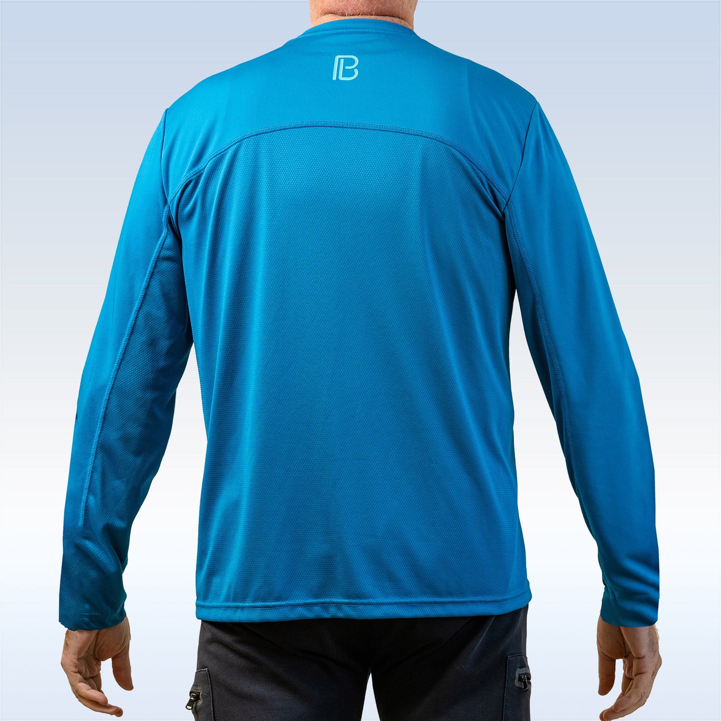 A person seen from behind wears a PB Men's Mykonos Blue Long Sleeve Tee by Pickleball Bella, featuring a small white stylized “B” logo on the upper back. The moisture-wicking shirt is paired with dark pants, set against a light gradient background.