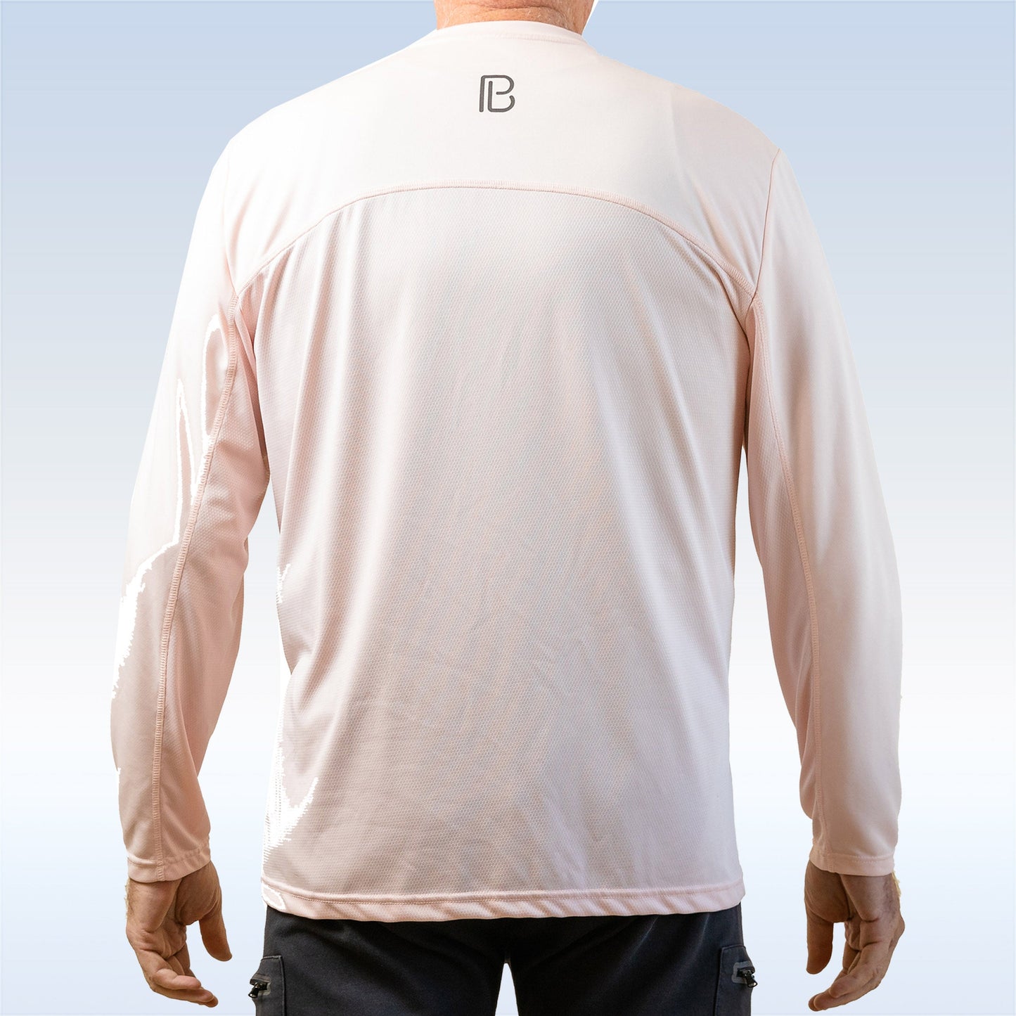 A person is seen from behind wearing the Pickleball Bella PB Men's Gossamer Pink Long Sleeve Tee, featuring a small black logo on the upper back. The moisture-wicking fabric and blue-to-white background evoke comfort and style.