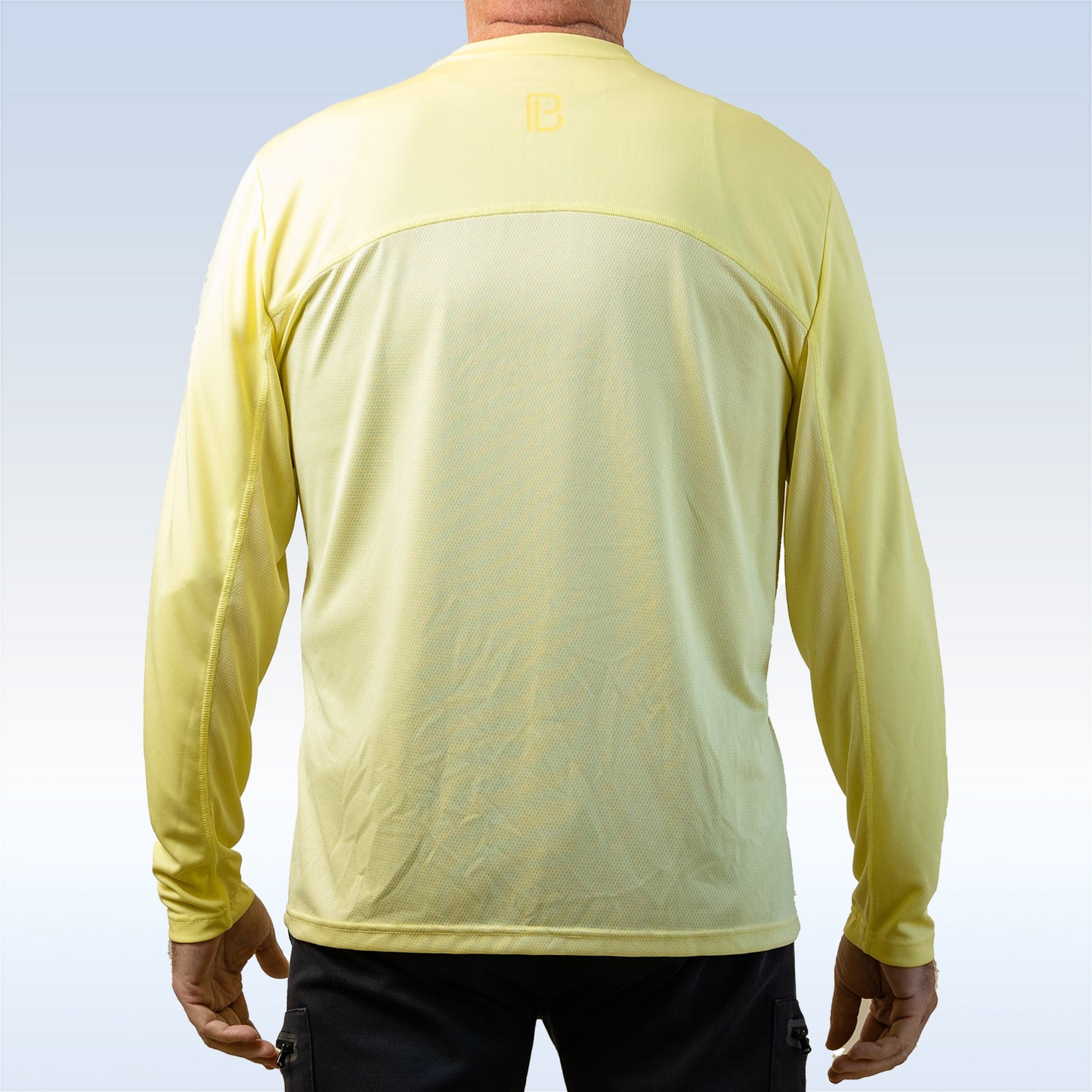 A man faces away, wearing a Pickleball Bella PB Men's Elfin Yellow Long Sleeve Tee with SPF 30+ and black pants. The shirt features a subtle logo near the neck and visible back seams. Background is a blue-to-white gradient.