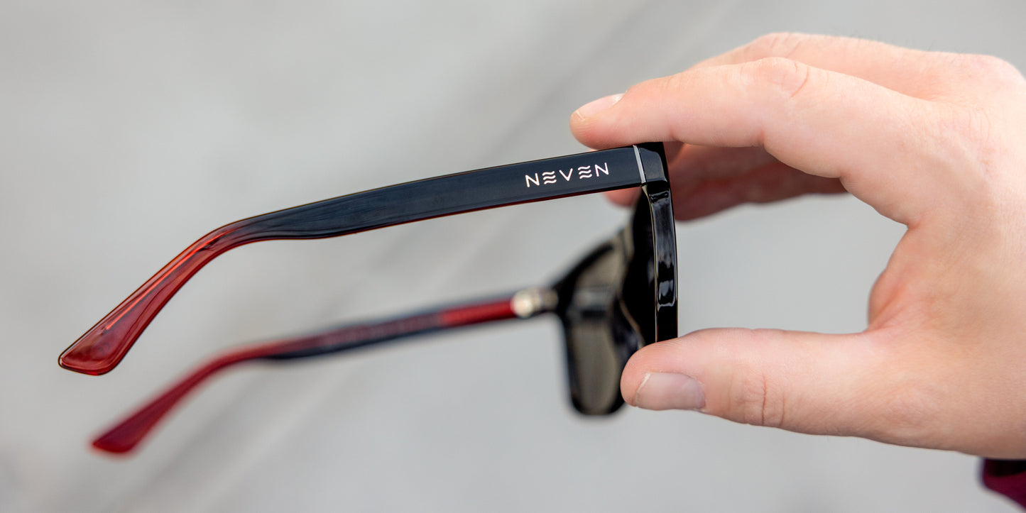A hand holds a pair of black rectangular frame Ignite glasses with red inner arms from Neven Eyewear. The white NEVEN logo appears on the temple arm. The background is a blurred gray.