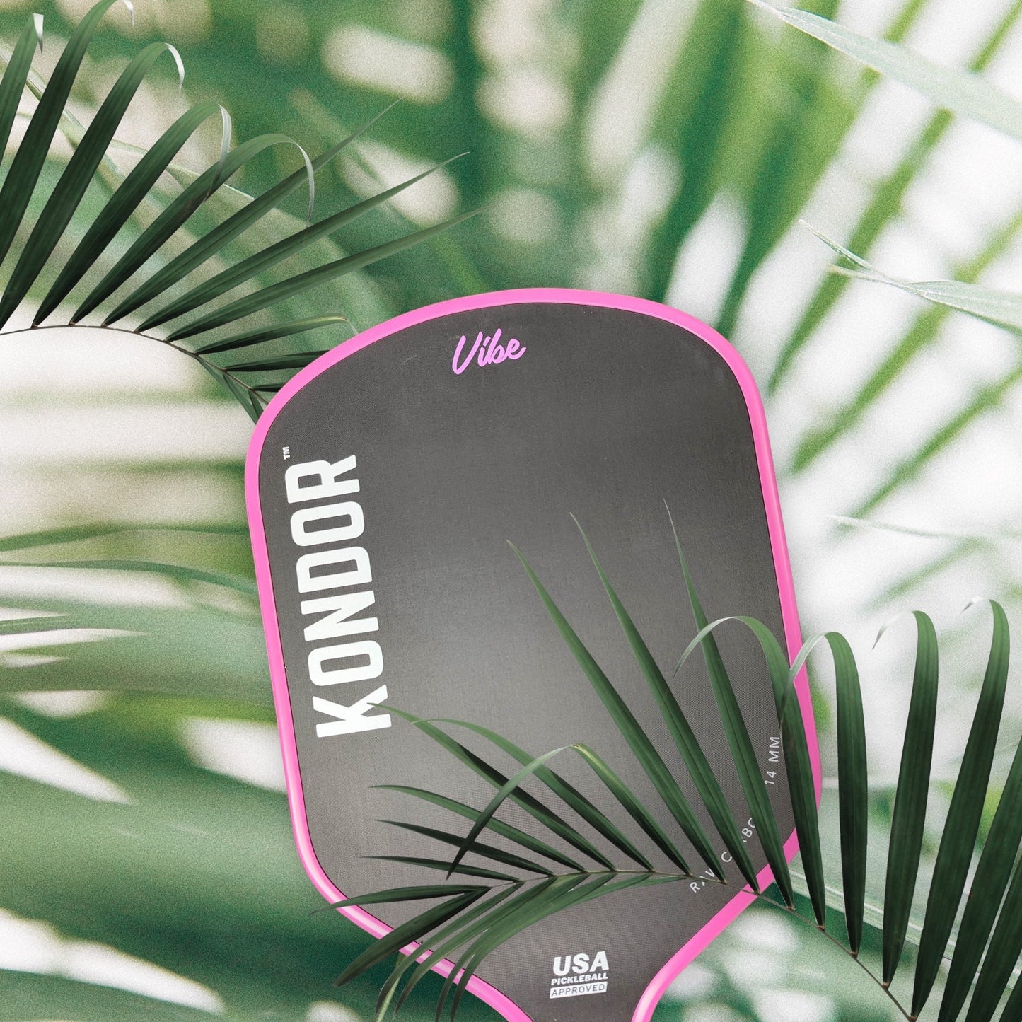 A black and pink Vibe Pro Power - TROPIXX SERIES pickleball paddle by KONDOR PICKLEBALL rests among palm leaves, sunlight casting tropical shadows. "USA Approved" is printed in white text at the bottom of the paddle.