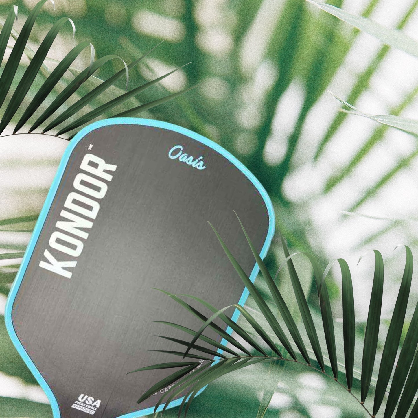 A gray KONDOR PICKLEBALL Oasis Pro Power - TROPIXX Series paddle with blue trim and logo is partially visible among lush green palm fronds, the softly blurred background highlighting both the paddle and tropical setting.