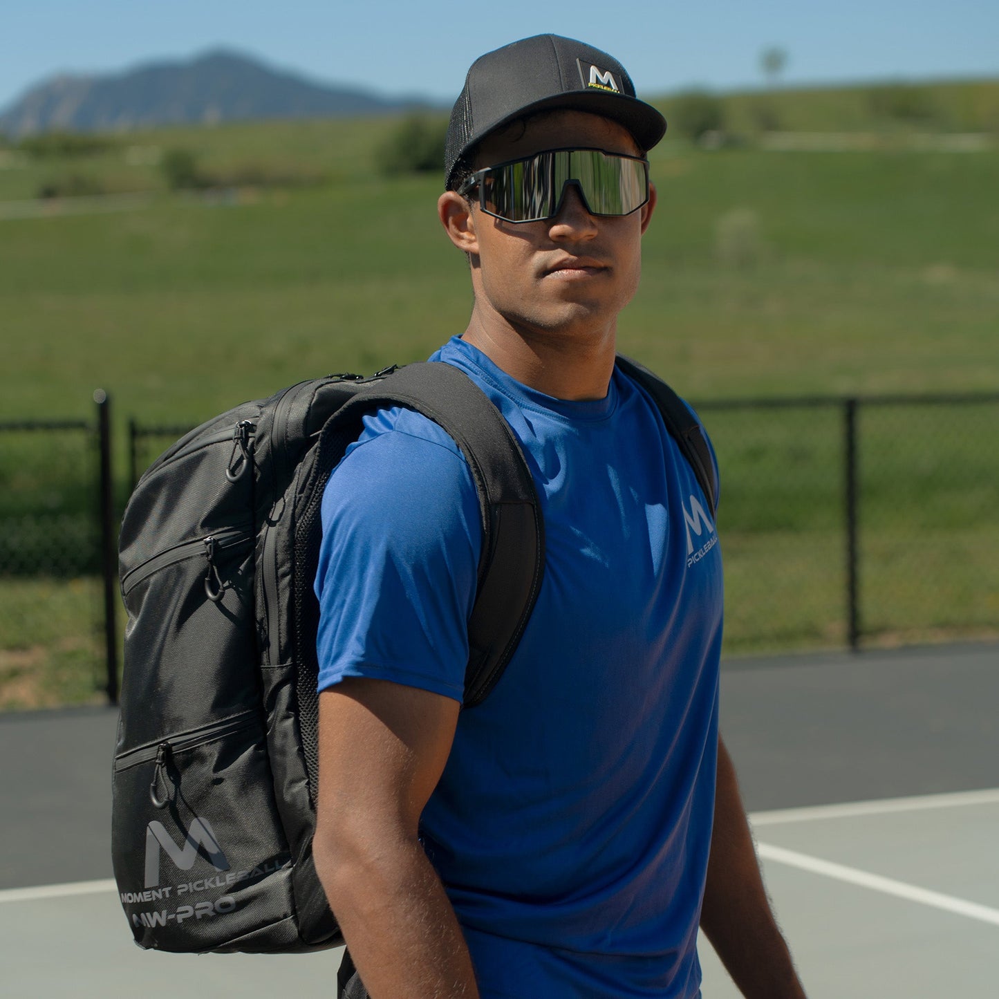 A man stands on an outdoor court with the Moment Pickleball MW-PRO Bag / Travel Bag (Black/Silver) over one shoulder. He wears a blue t-shirt, black cap, and reflective sunglasses, with a metal fence and mountains in the background.