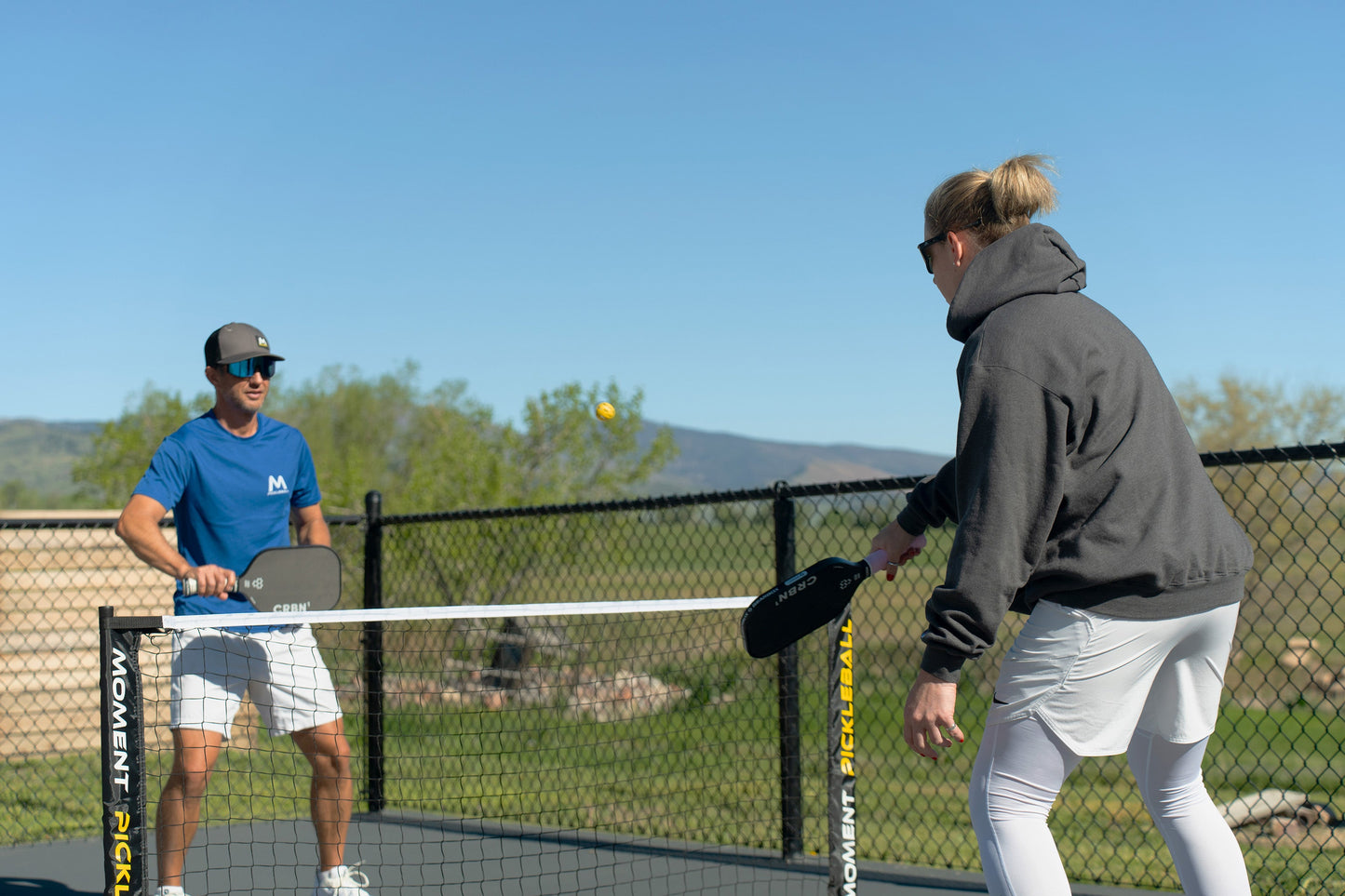Two people play pickleball outdoors with a black net, using Moment Pickleblasters 2-Color Mini Plastic Practice Balls by Moment Pickleball. A yellow ball is in midair, with trees and mountains as a scenic game backdrop.