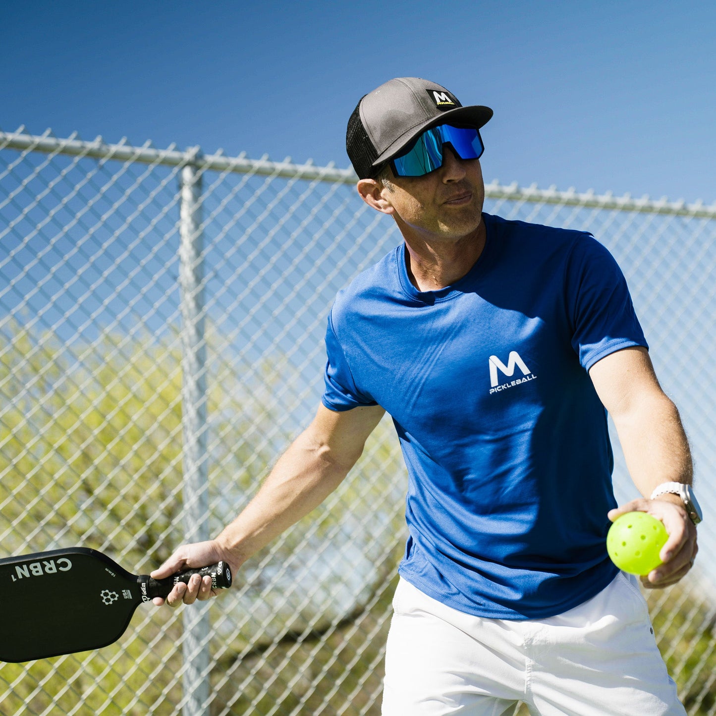 A man in a blue shirt and white shorts, wearing Moment Pickleball's Pro-Guard Glasses Set with Blue Mirrored Lens and a cap, holds a pickleball paddle and yellow ball outdoors by a chain-link fence, ready to serve under the clear blue sky.